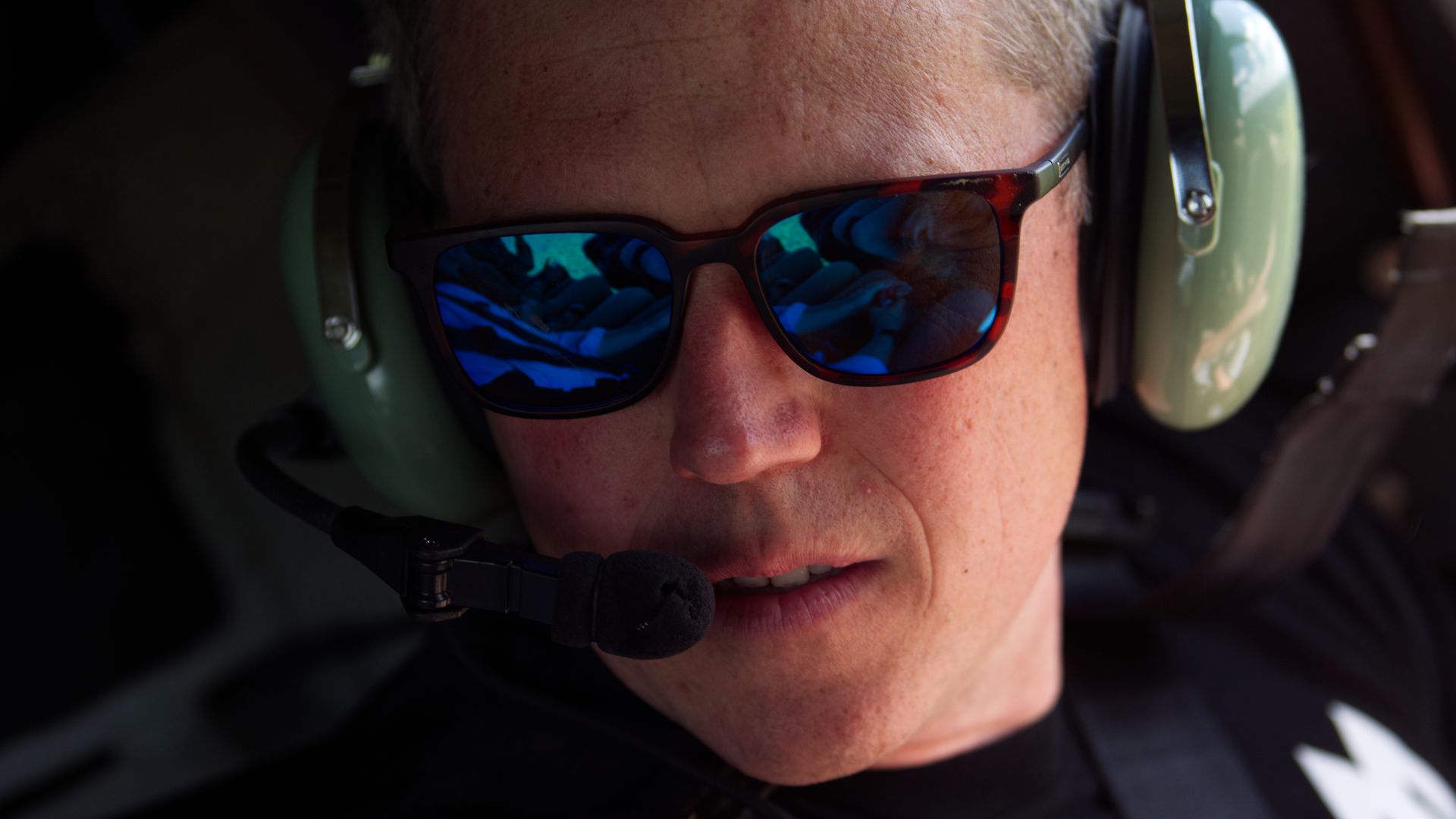Close-up of a man wearing blue-tinted sunglasses, green aviation headset with microphone, and a black shirt, with reflections visible in the sunglasses lenses.