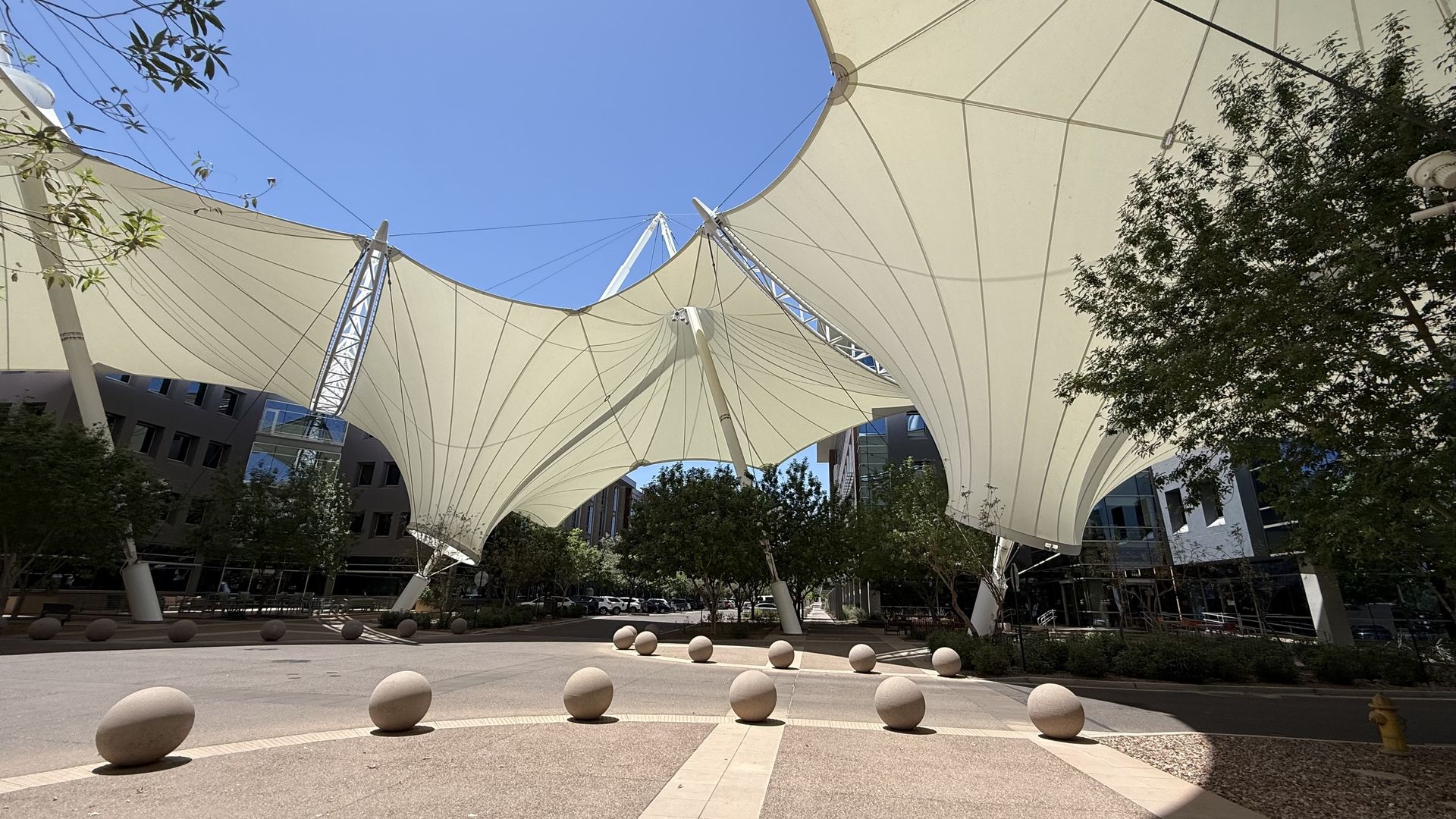 Wide courtyard with white tensile fabric canopies on tall masts and cables, forming curved sails. Round concrete bollards line the plaza, with trees and modern buildings under a blue sky.