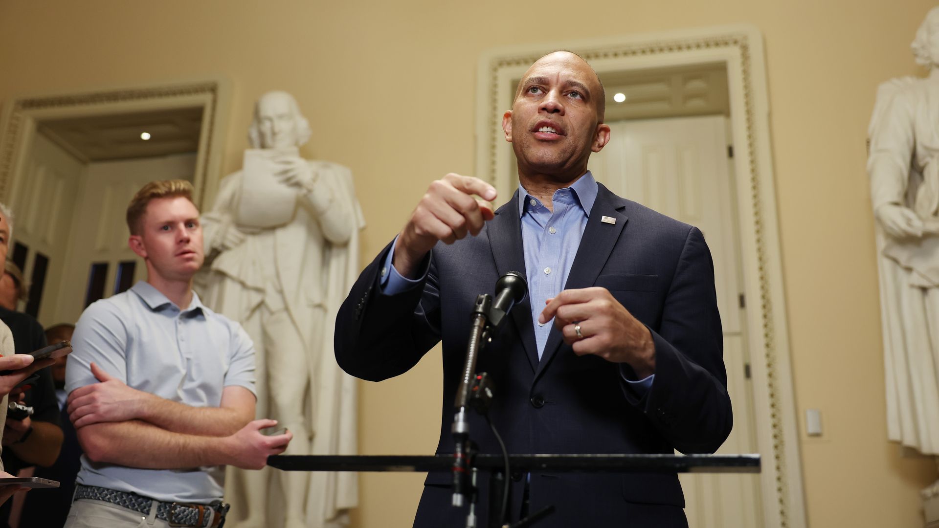 Hakeem Jeffries, wearing a blue suit and gesturing with both hands as he stands behind a podium and in front of a white wall and statues.
