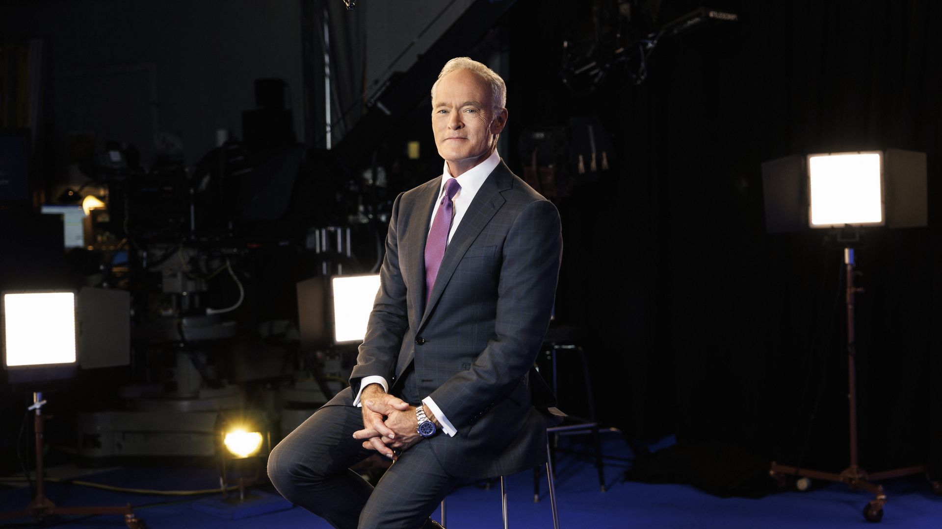 "60 Minutes" correspondent Scott Pelley, an older man with gray hair, wearing a navy suit, stares into the camera while sitting on a chair in a dark studio that features spotlights."