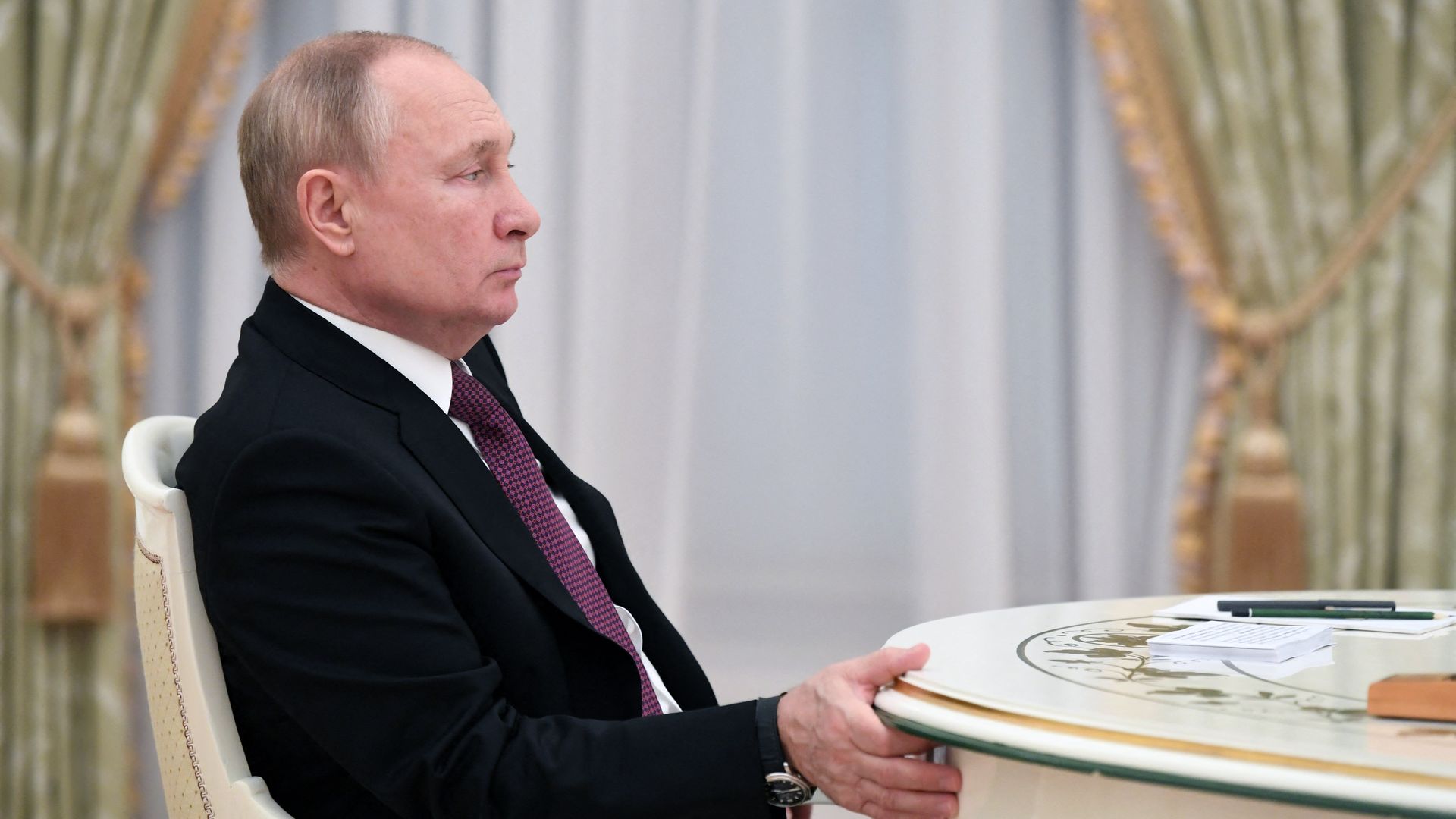 Vladimir Putin sits at a table with his right hand on the edge