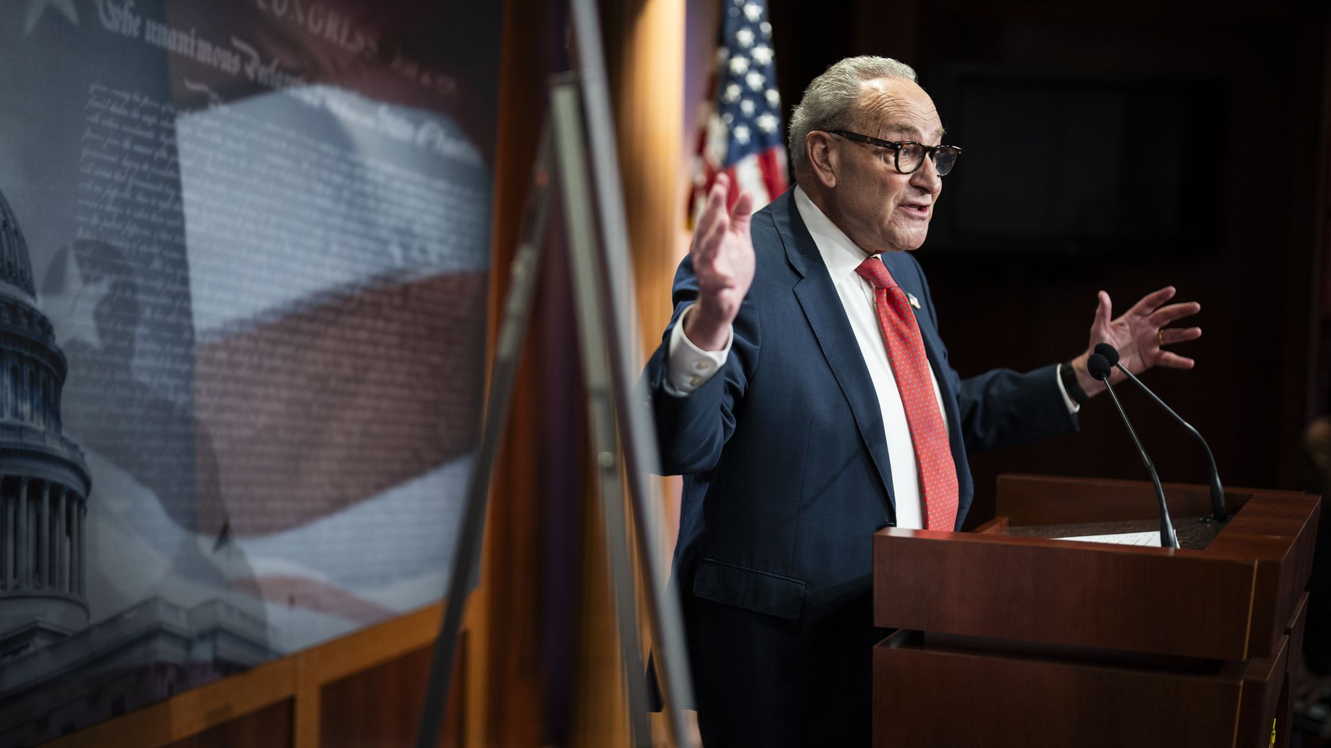 Chuck Schumer, wearing a blue suit and standing behind a wooden podium in front of an American flag and an American flag mural with his arms raised.
