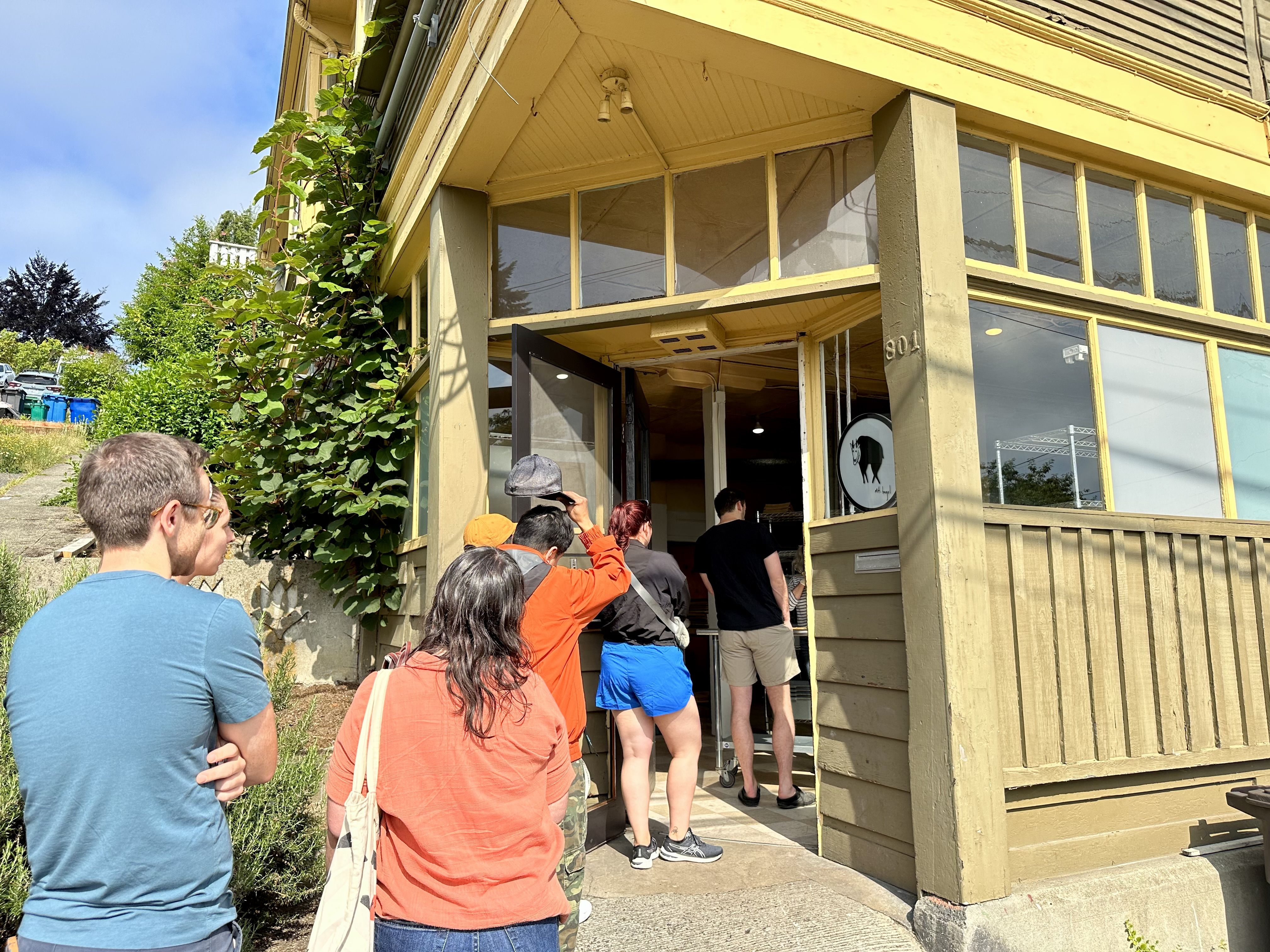 People stand outside in a line waiting to get into Mt. Bagel, which is a corner shop painted yellow.