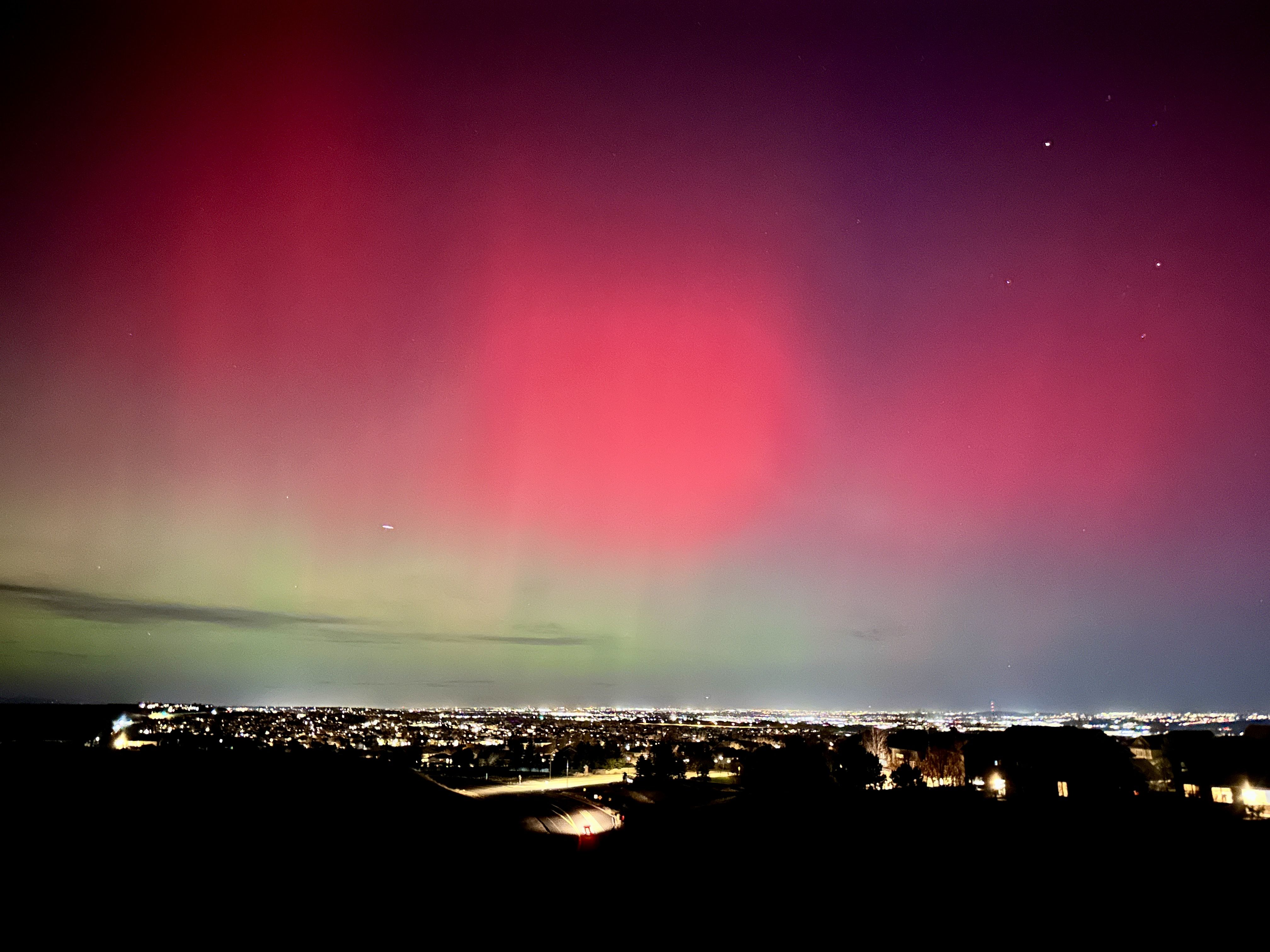 City lights under a night sky with red and green northern lights, stars visible, and a dark foreground of houses and trees.