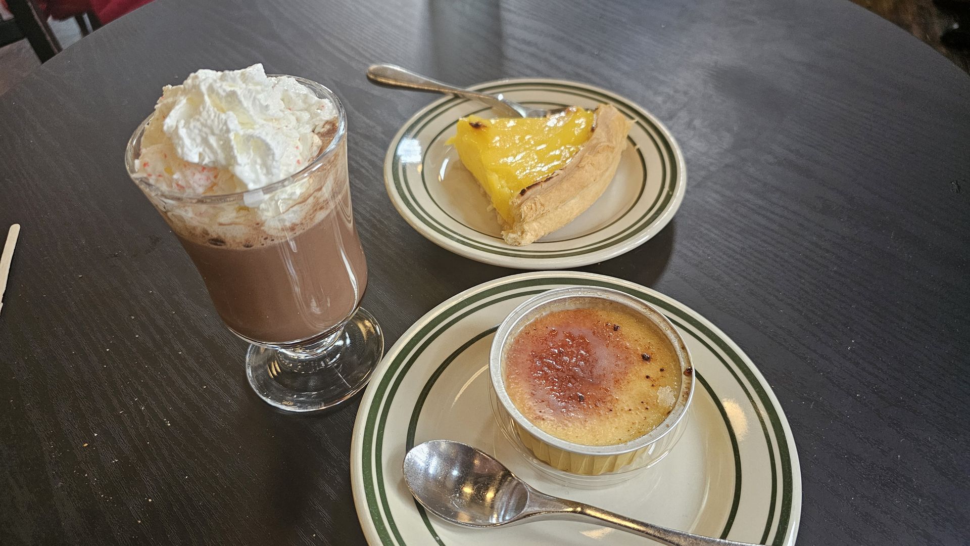 Glass of hot chocolate with whipped cream, slice of yellow fruit tart on a plate with spoon, and crème brûlée in a ramekin with a spoon on a dark wooden table.