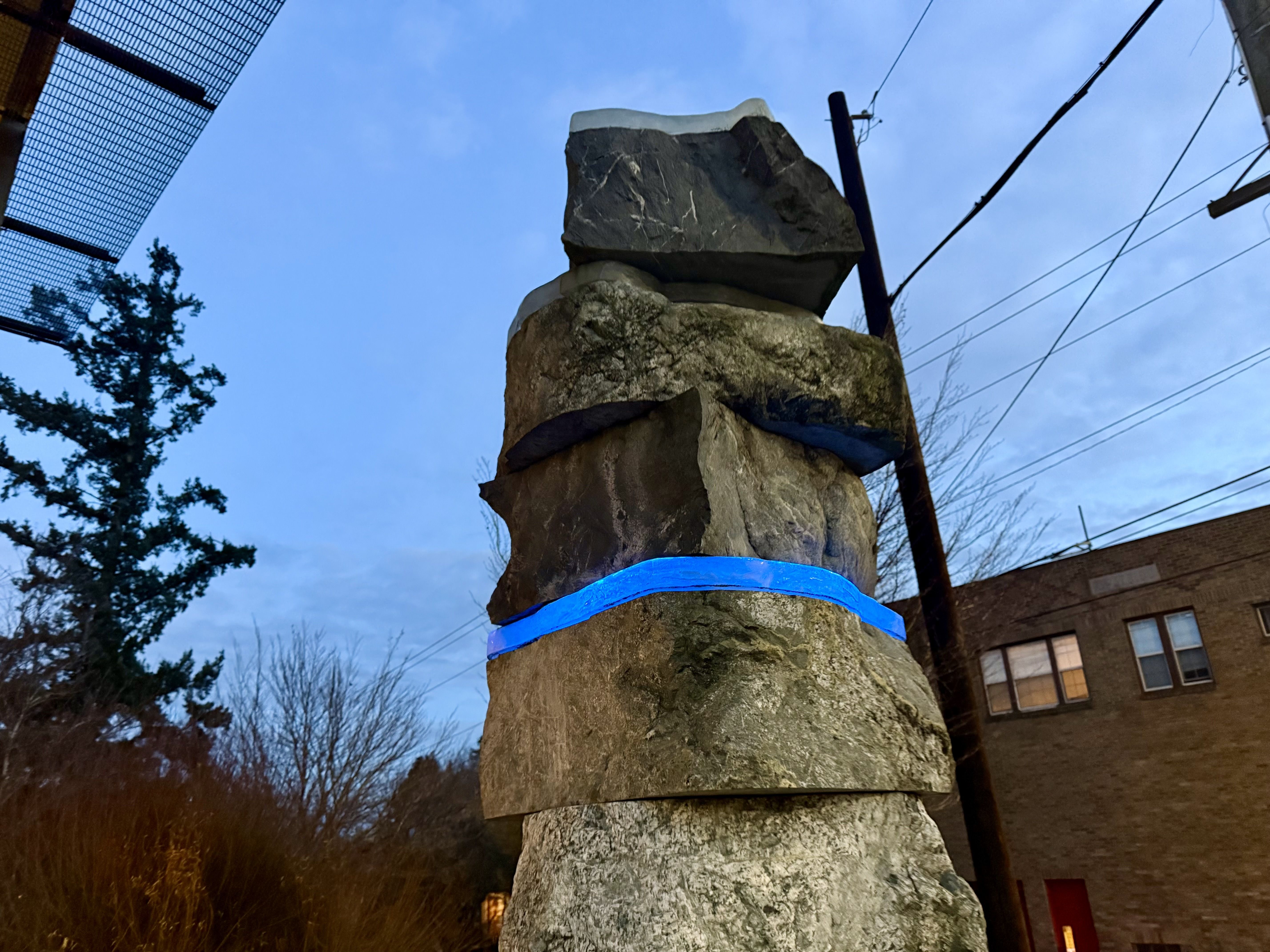 Stone pillar sculpture with a glowing blue band around its middle, set outdoors under a blue early evening sky near trees, power lines, and a brick building with lit windows.