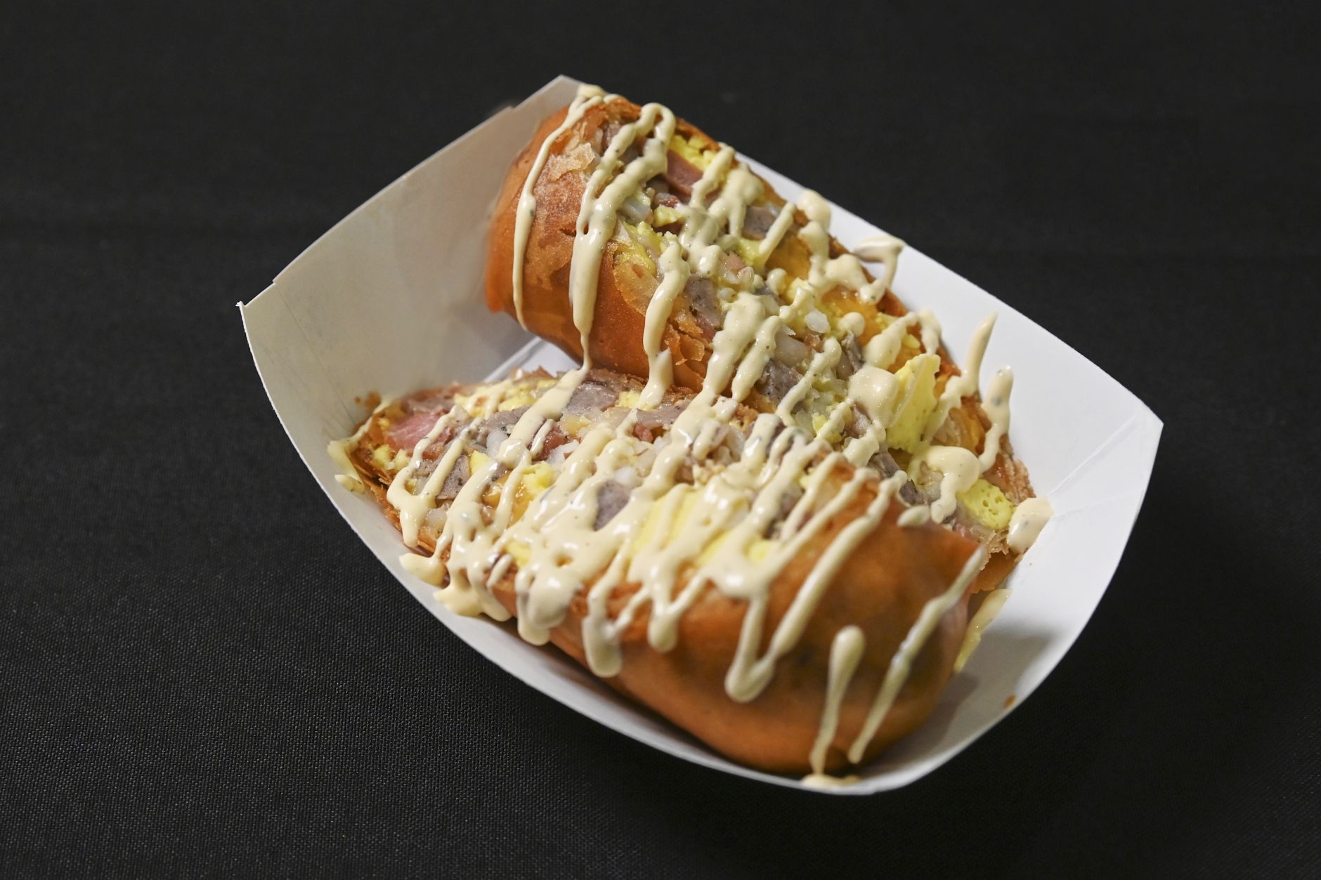 An egg roll cut in half in a white takeout tray, topped with ranch, and a zigzag of creamy mayo on a dark background.