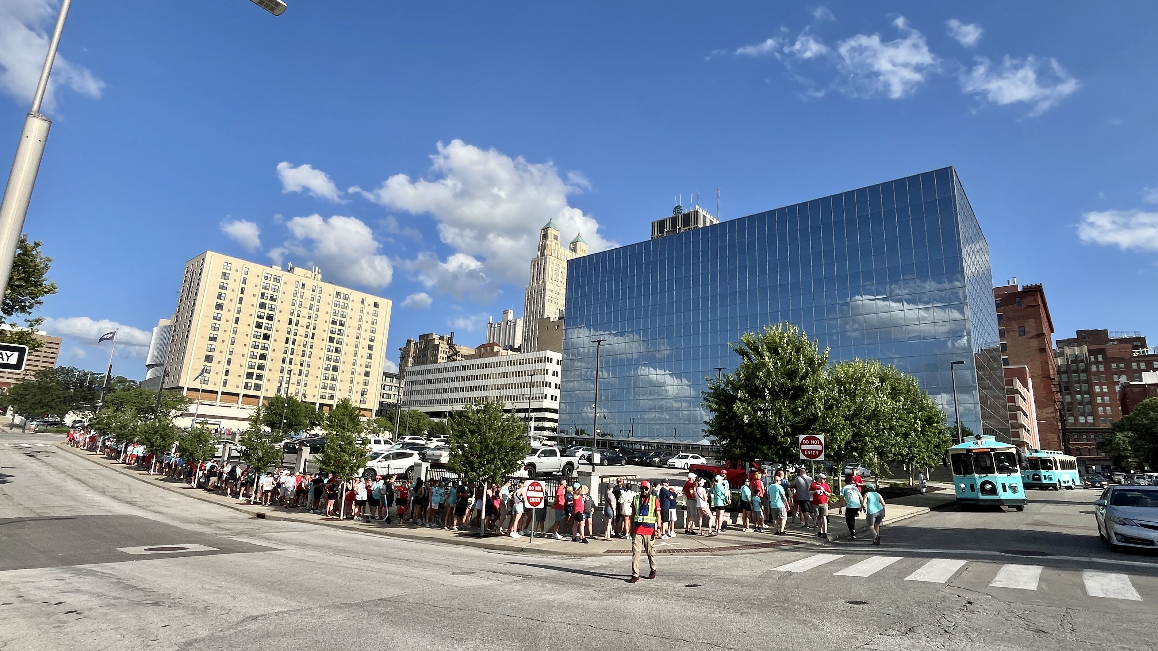 City street scene with a long line of KC Current fans waiting along the sidewalk for a gameday shuttle. A reflective blue glass office building, a beige apartment block, and a turquoise bus under a bright blue sky at the corner of 7th and Baltimore downtown Kansas City.
