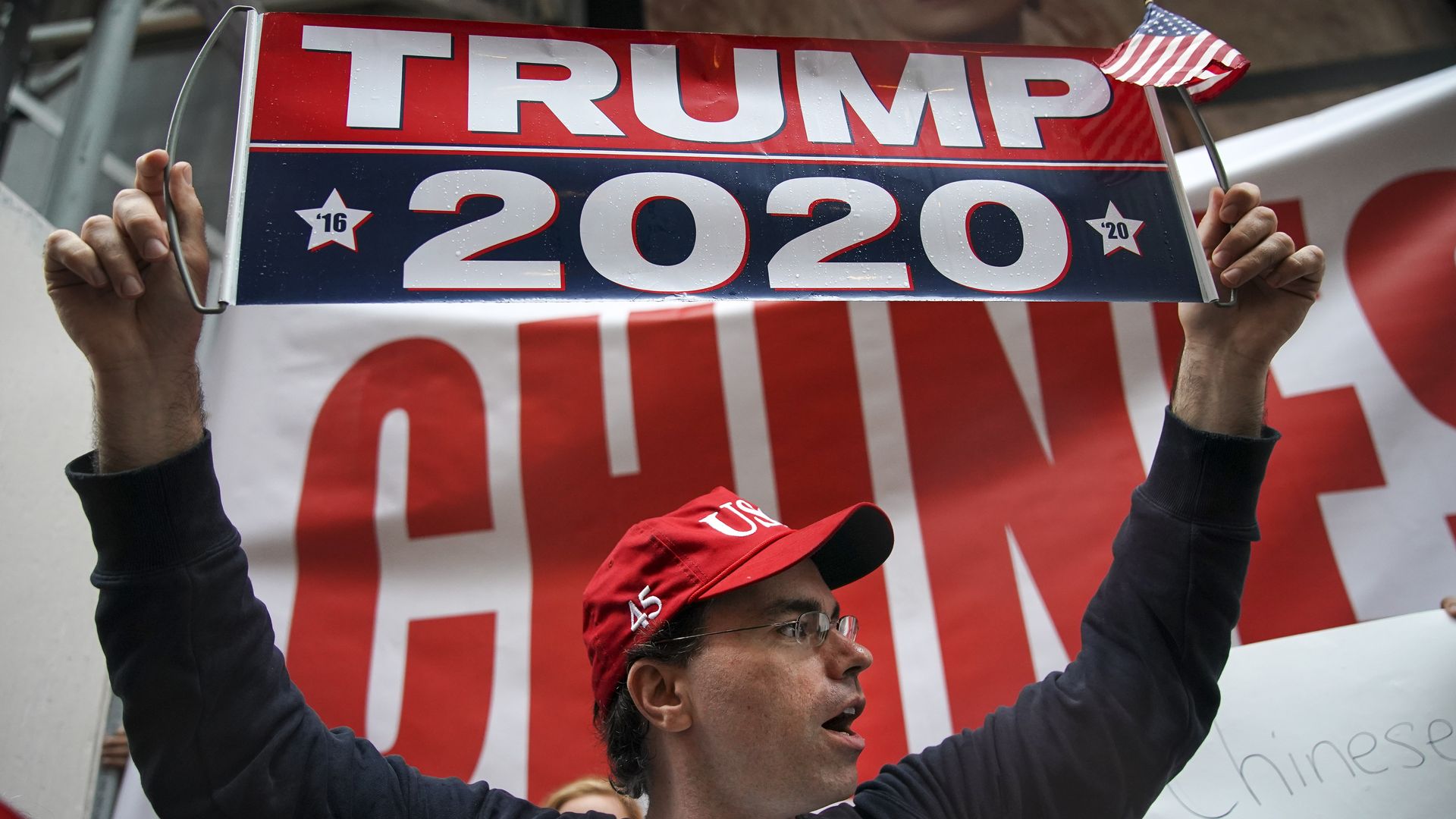 In this image, a man holds up a banner that reads "Trump 2020"