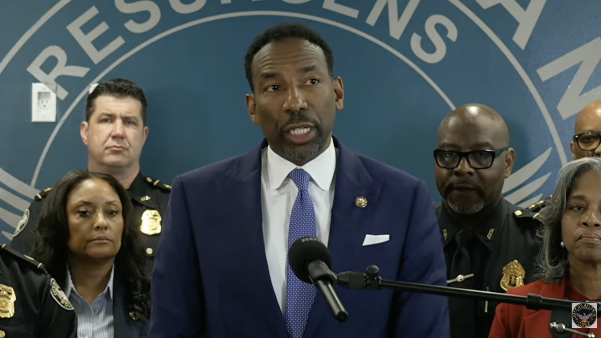 A man in a navy suit and purple tie speaks at a microphone at a press conference, flanked by uniformed officers and officials, with a blue wall and partial circular emblem in the background.