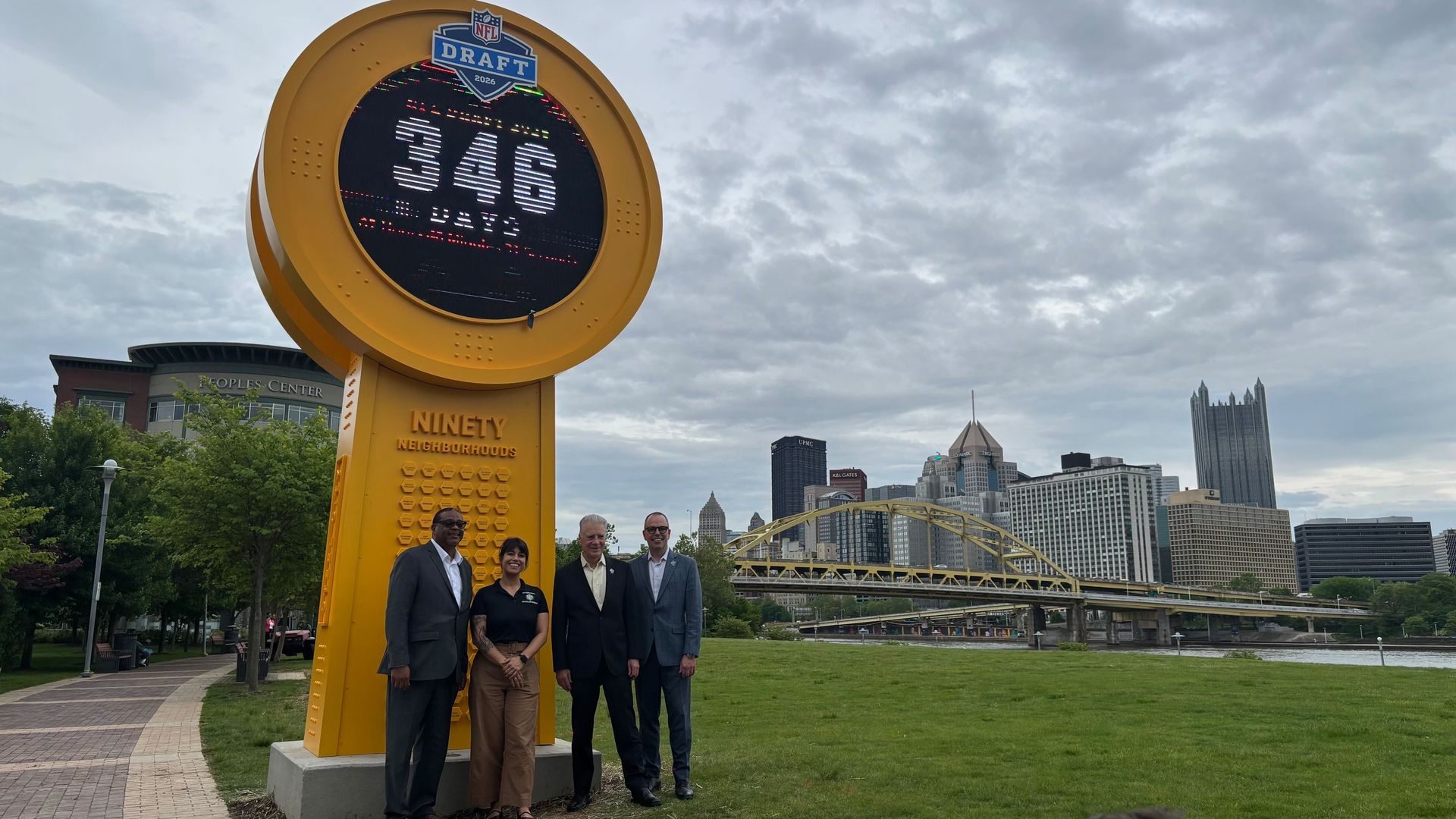 Four people stand in front of a large sign that says "346" and is counting down to the NFL Draft in 2026. The sign is in front of the Pittsburgh skyline on a cloudy day.