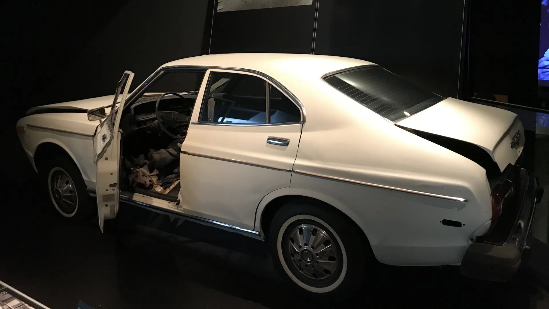 White 1970s-style four-door car with open driver’s door and visible interior, set against a dark background, with the hood partially buckled and the trunk partially open. 