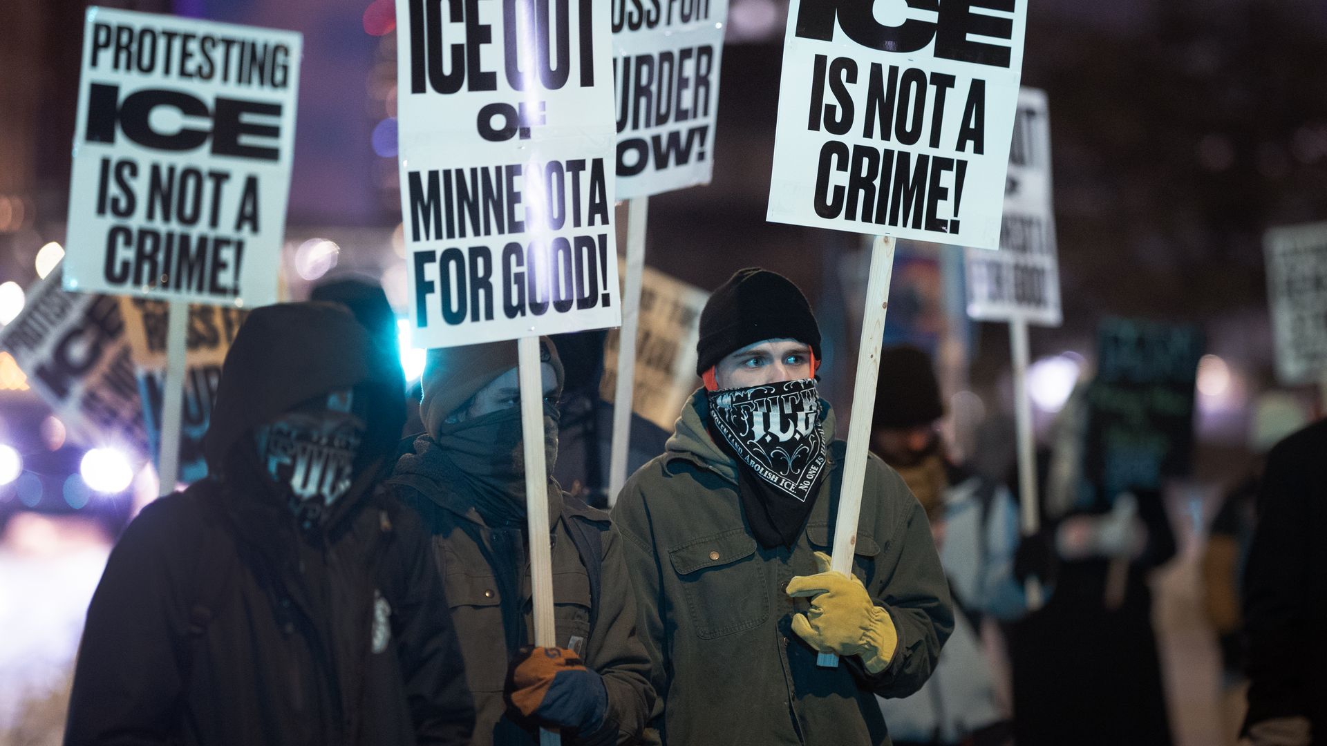 Protesters wearing winter clothes and face coverings hold signs at night saying "Protesting ICE is not a crime!" and "Ice out of Minnesota for good!"