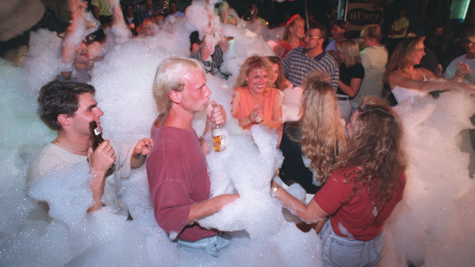 Crowded foam party at night with dozens of people dancing and laughing amid thick white bubbles. Many hold drinks; banners and club signs loom in the background.