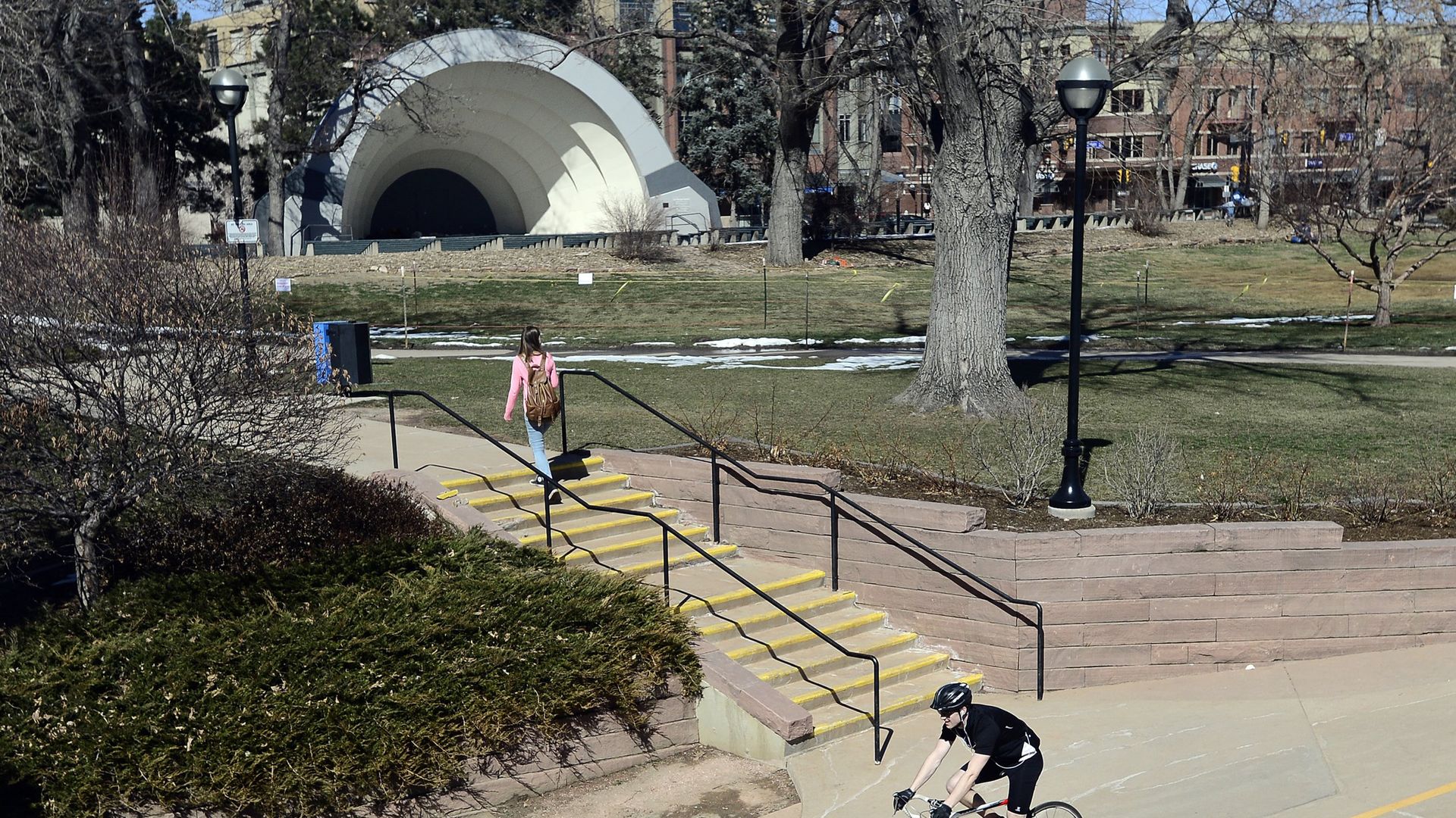 A cyclist in black rides along a curved concrete bike path in a park. A woman in a pink jacket climbs yellow-edged stairs. In the background, a large white arched tunnel and bare trees.