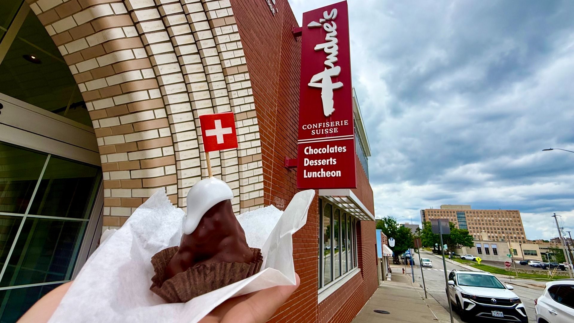 Hand holds a chocolate ice cream cone with a white flag topper showing a Swiss cross, in front of a brick building and a tall red Confiserie Suisse sign listing chocolates, desserts, luncheon; cloudy sky.