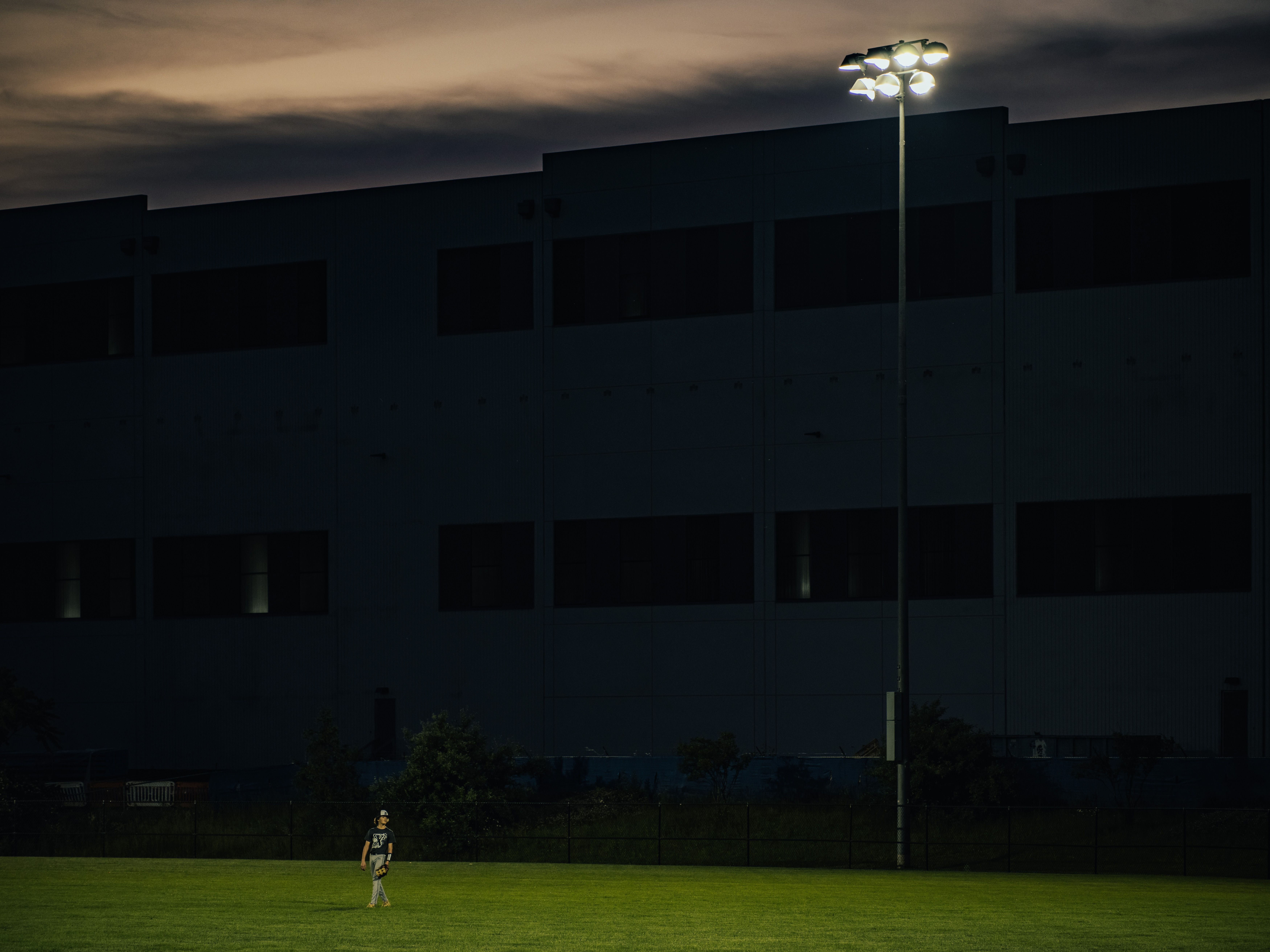 Night view of a dark, windowed building with a bright floodlight tower on the right. A person in sports gear stands on a green field at the bottom left under a cloudy, orange sky.