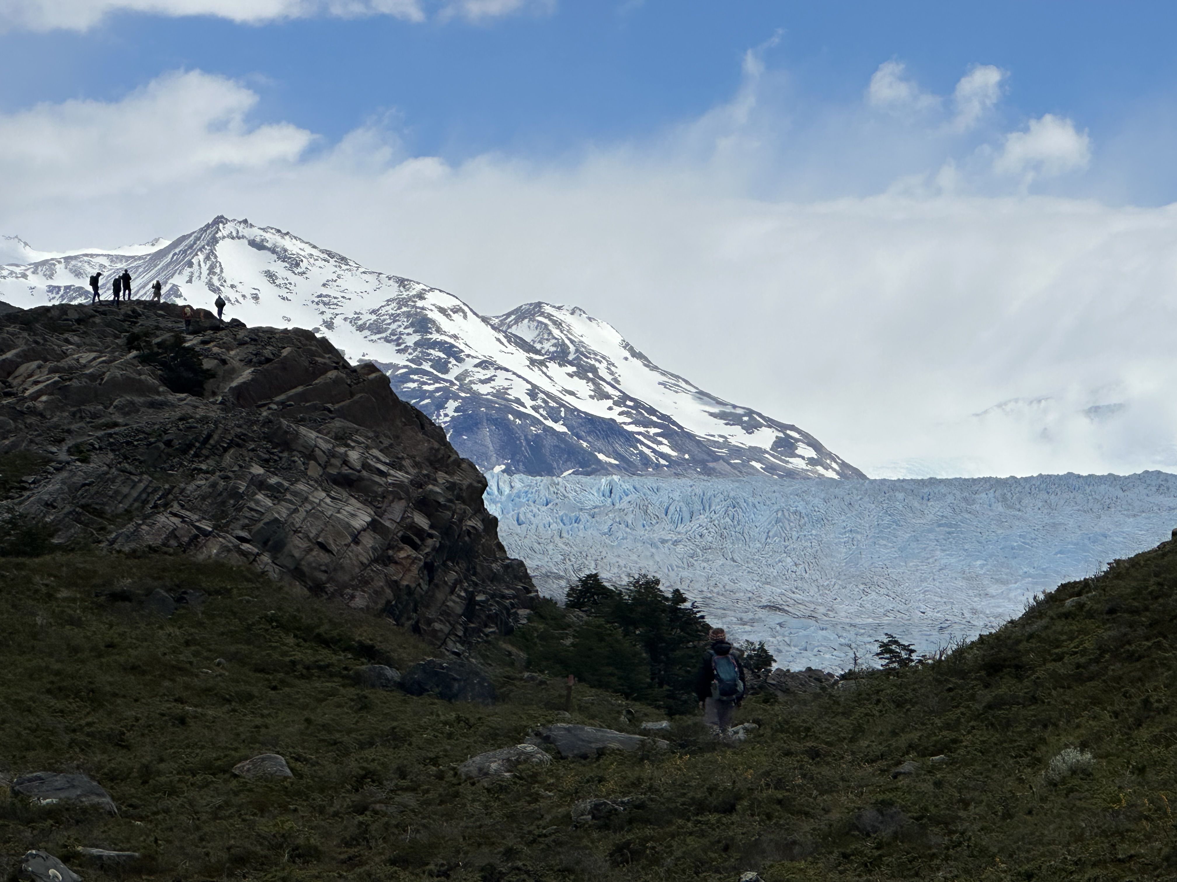 Hikers on rocky terrain with snow-capped mountains and a large blue glacier in the background under a partly cloudy sky.