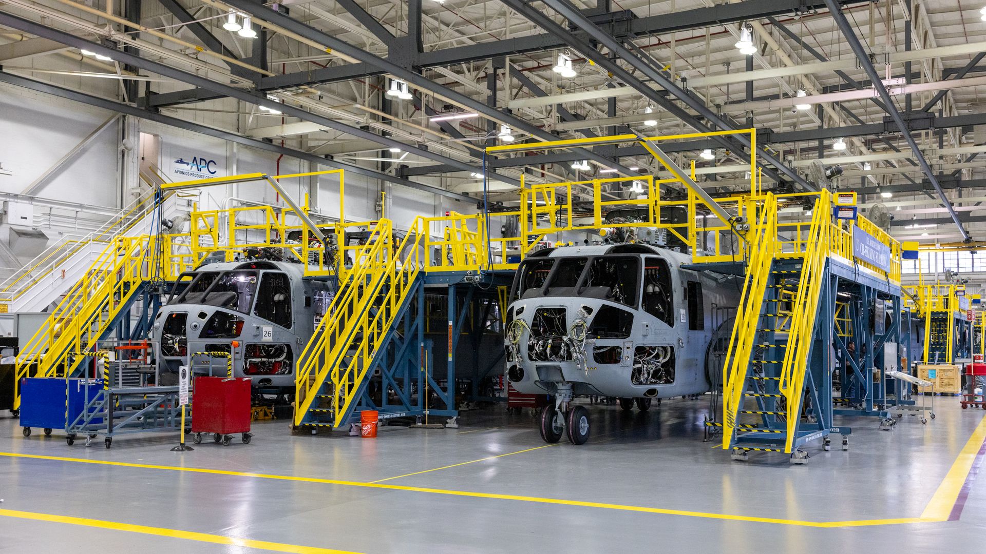Two unfinished gray helicopter fuselages in a brightly lit factory with yellow metal scaffolding and stairs surrounding them, industrial setting with tools and carts nearby.