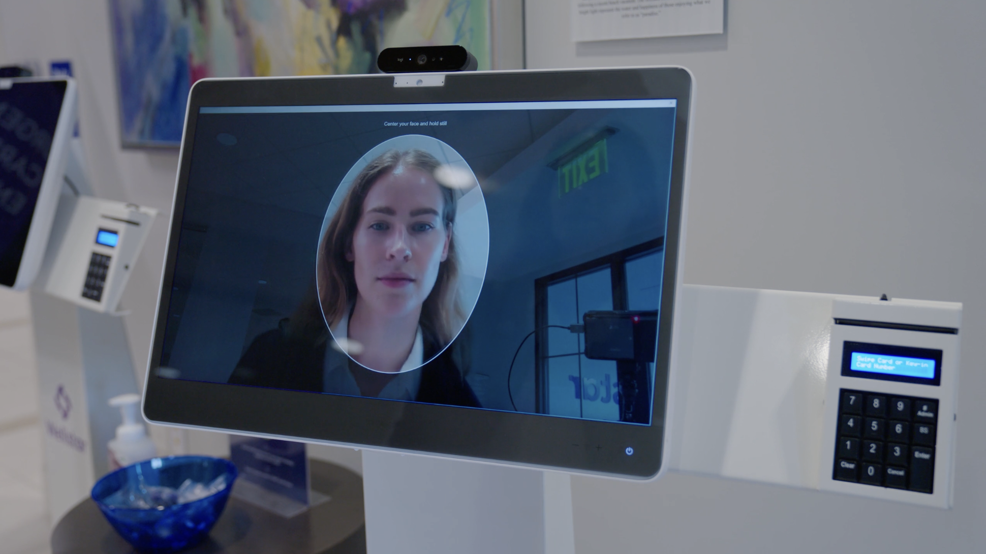 Close-up of a computer screen showing a woman's face inside a circle for facial recognition, with a keypad and card reader to the right, in an indoor setting with signs and equipment.