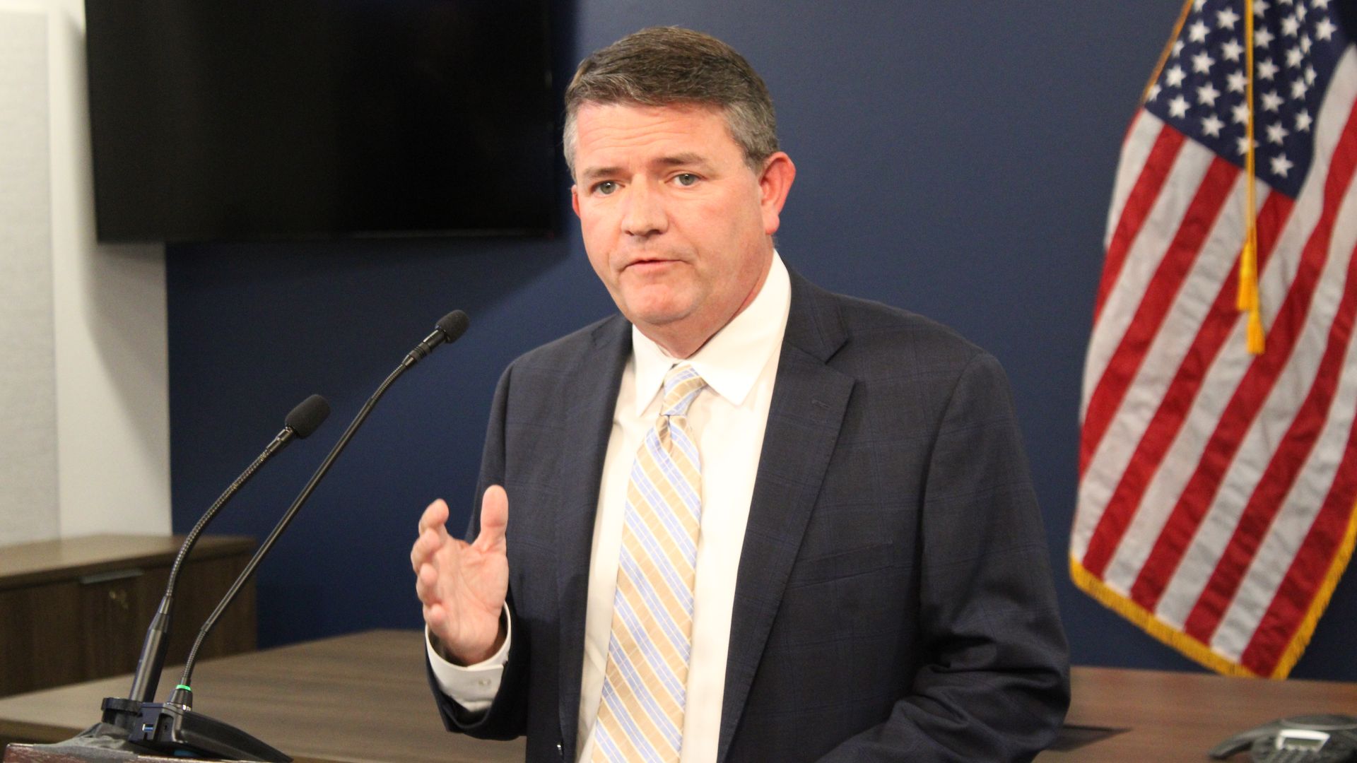 A man in a suit speaks at a lectern while gesturing with his right hand. 