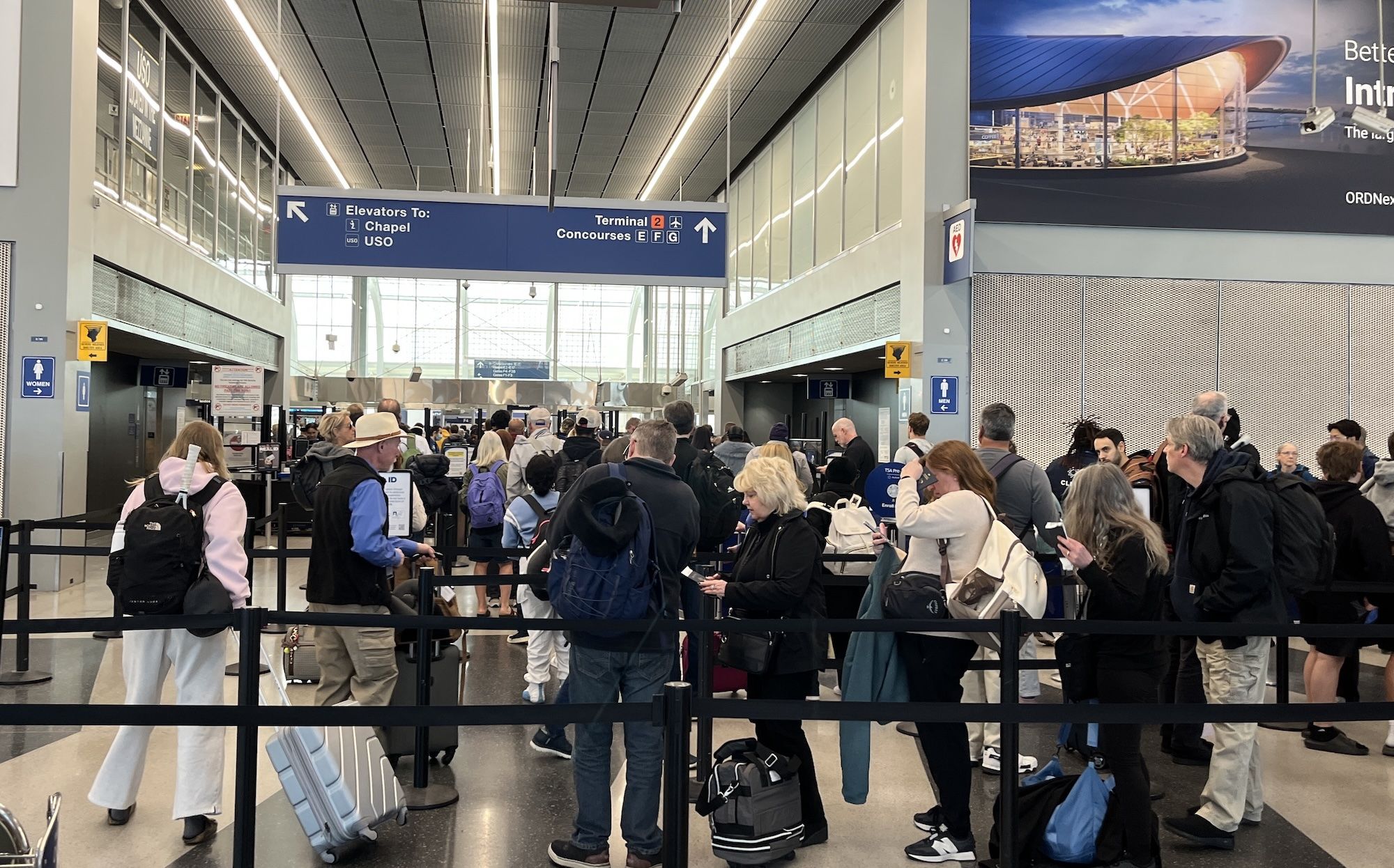 Busy airport terminal with a long queue of travelers and luggage. Blue overhead signs point to elevators, chapel, USO, and concourse; bright, modern interior.