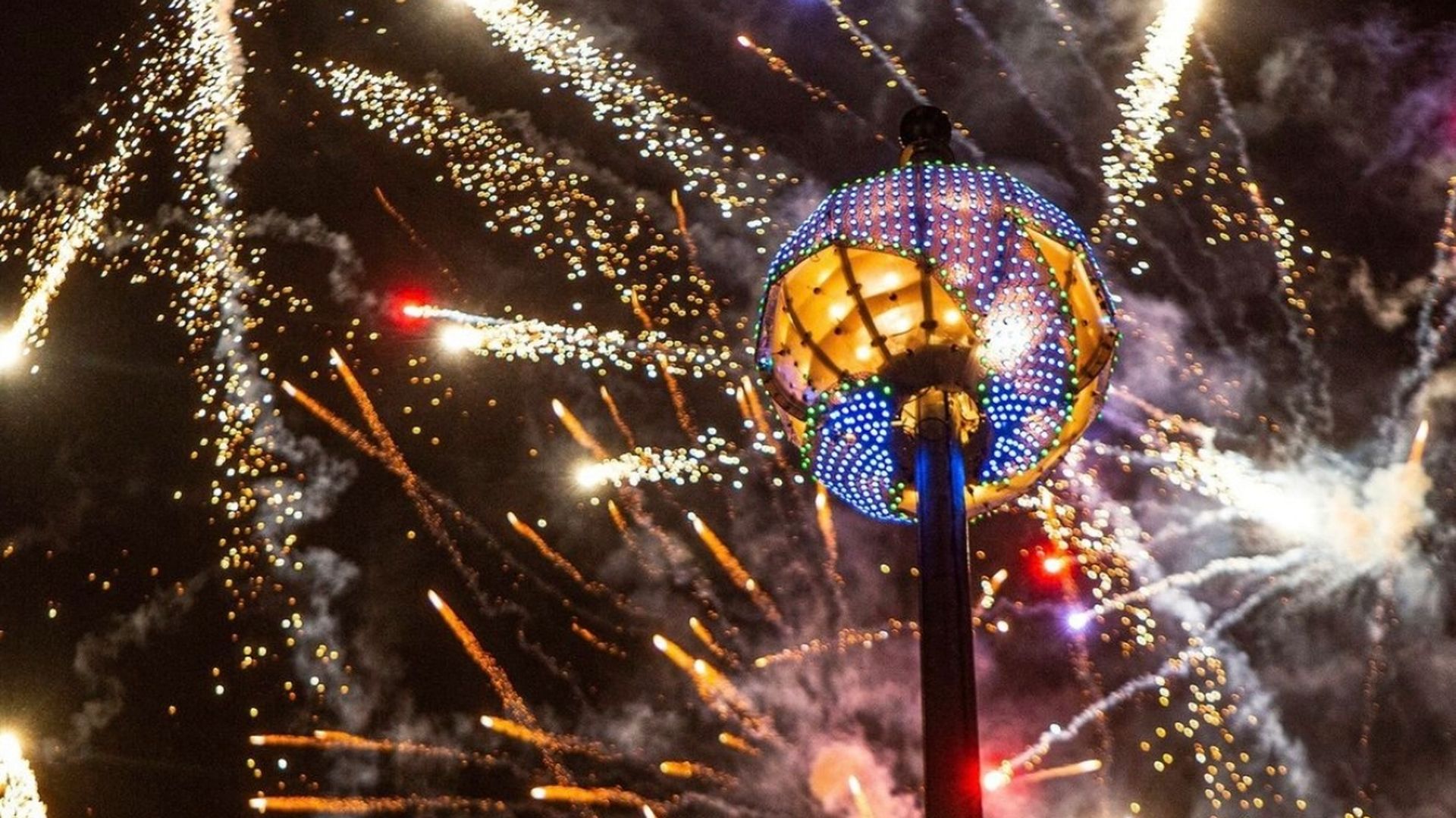 Night sky filled with colorful fireworks bursting around a tall pole topped with a large decorative, multi-colored illuminated orb light.