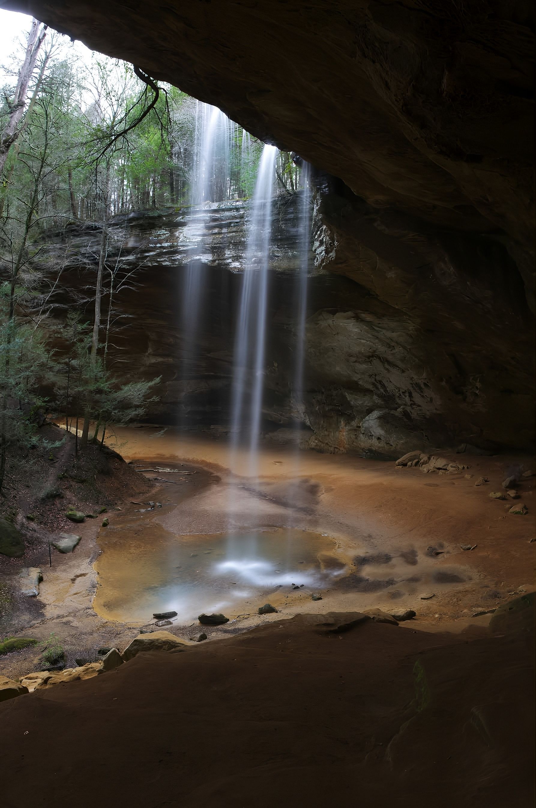 Under a dark rock overhang, two thin waterfalls spill from a cliff into a shallow, orange-brown pool. Surrounding trees and mossy rocks frame the tranquil scene.