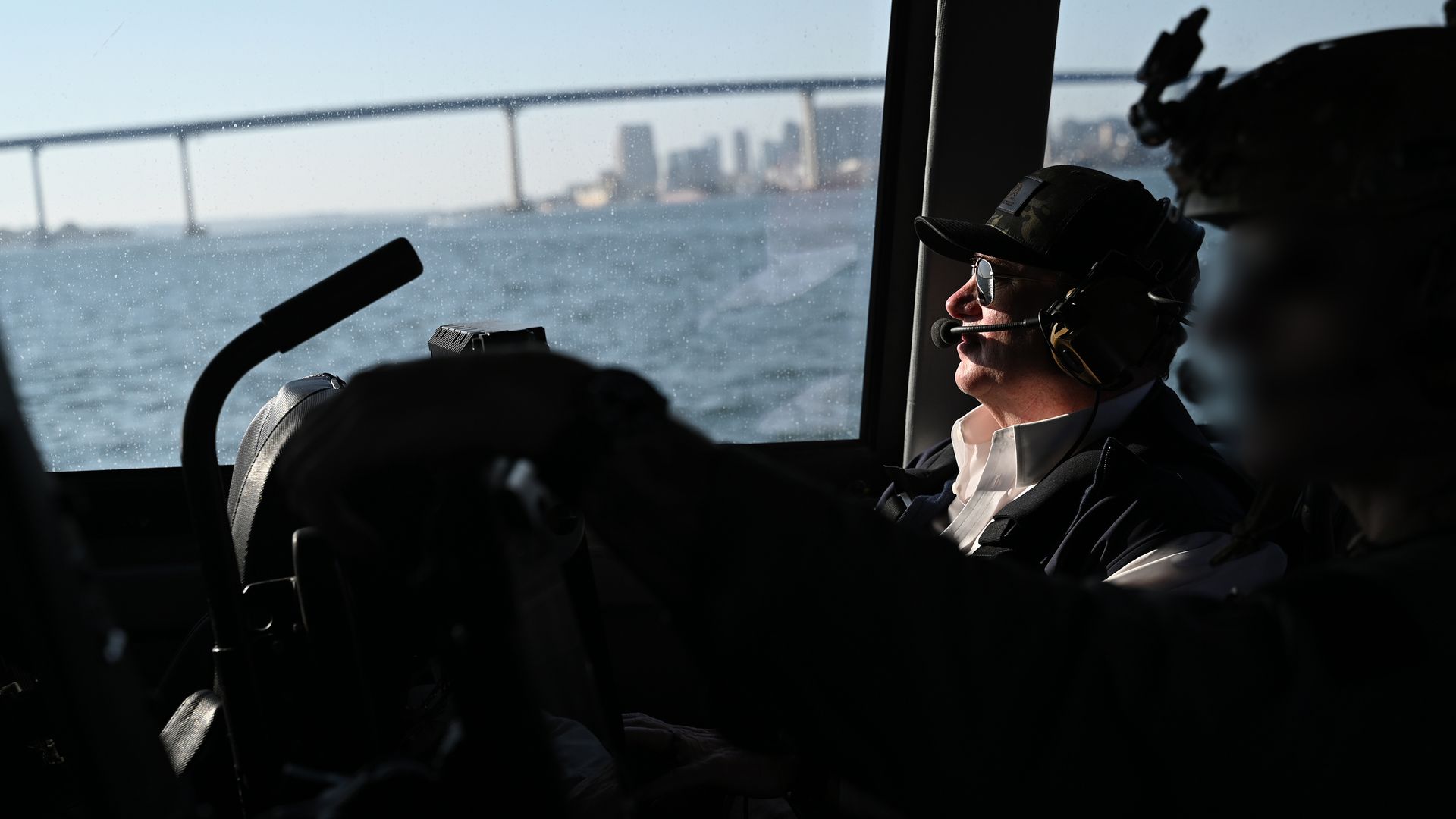 Man wearing a headset with microphone and sunglasses, sitting by a window inside a vehicle with water and a bridge visible outside in daylight.