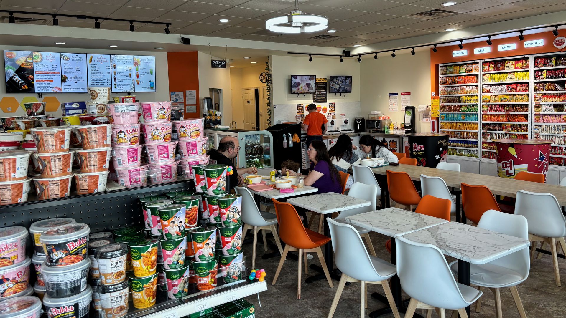 Bright casual market eatery with stacks of instant noodle cups in the foreground, a snack shelf along the right, and white-and-orange chairs around marble tables; a worker in orange stands at the counter.