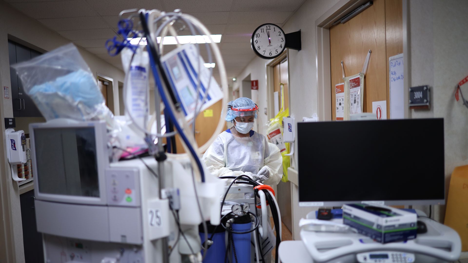 A health care worker in a white gown surrounded by medical equipment in a hospital ICU.