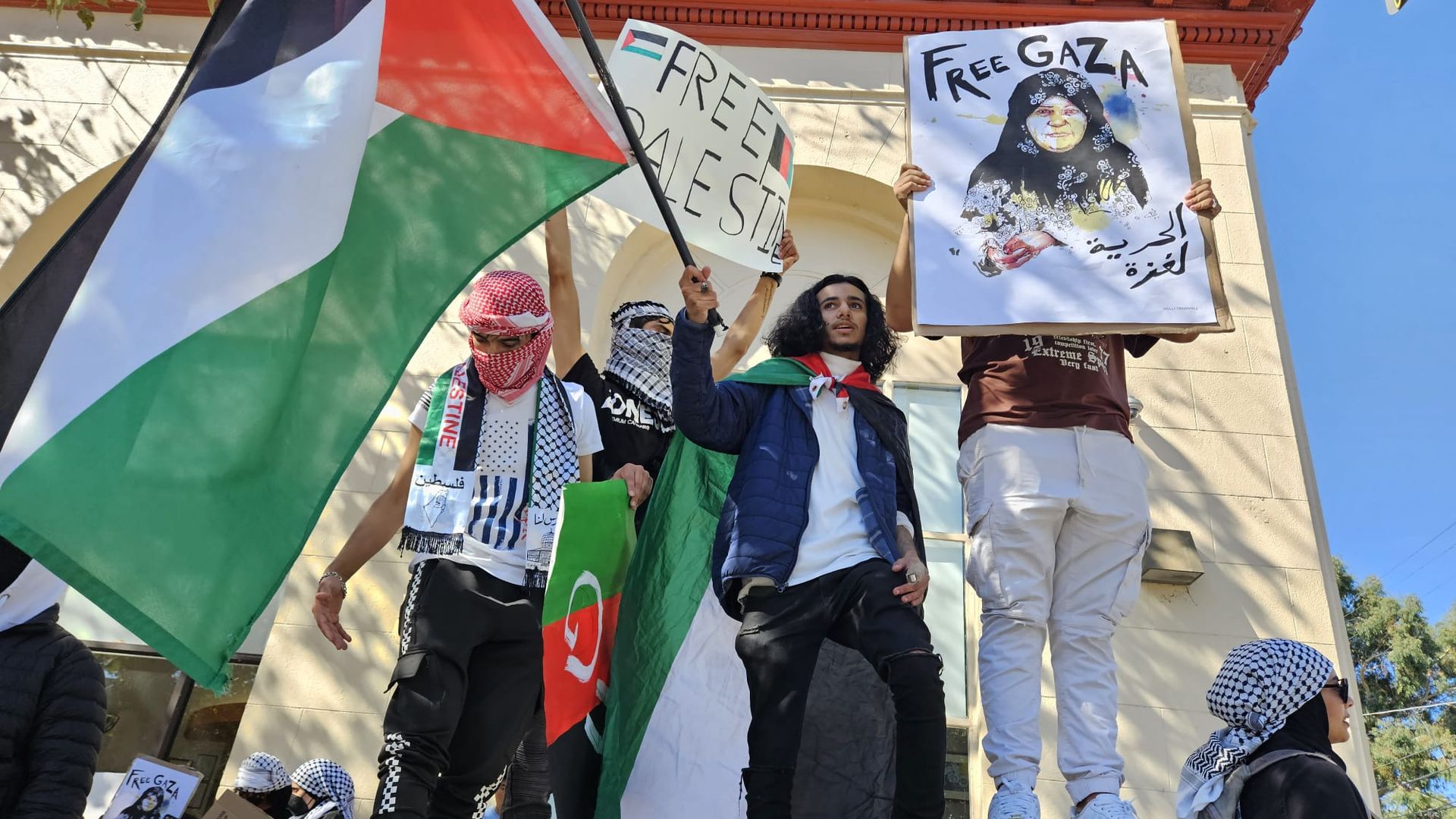 High school students stand on a platform raising the Palestinian flag and "Free Gaza" signs