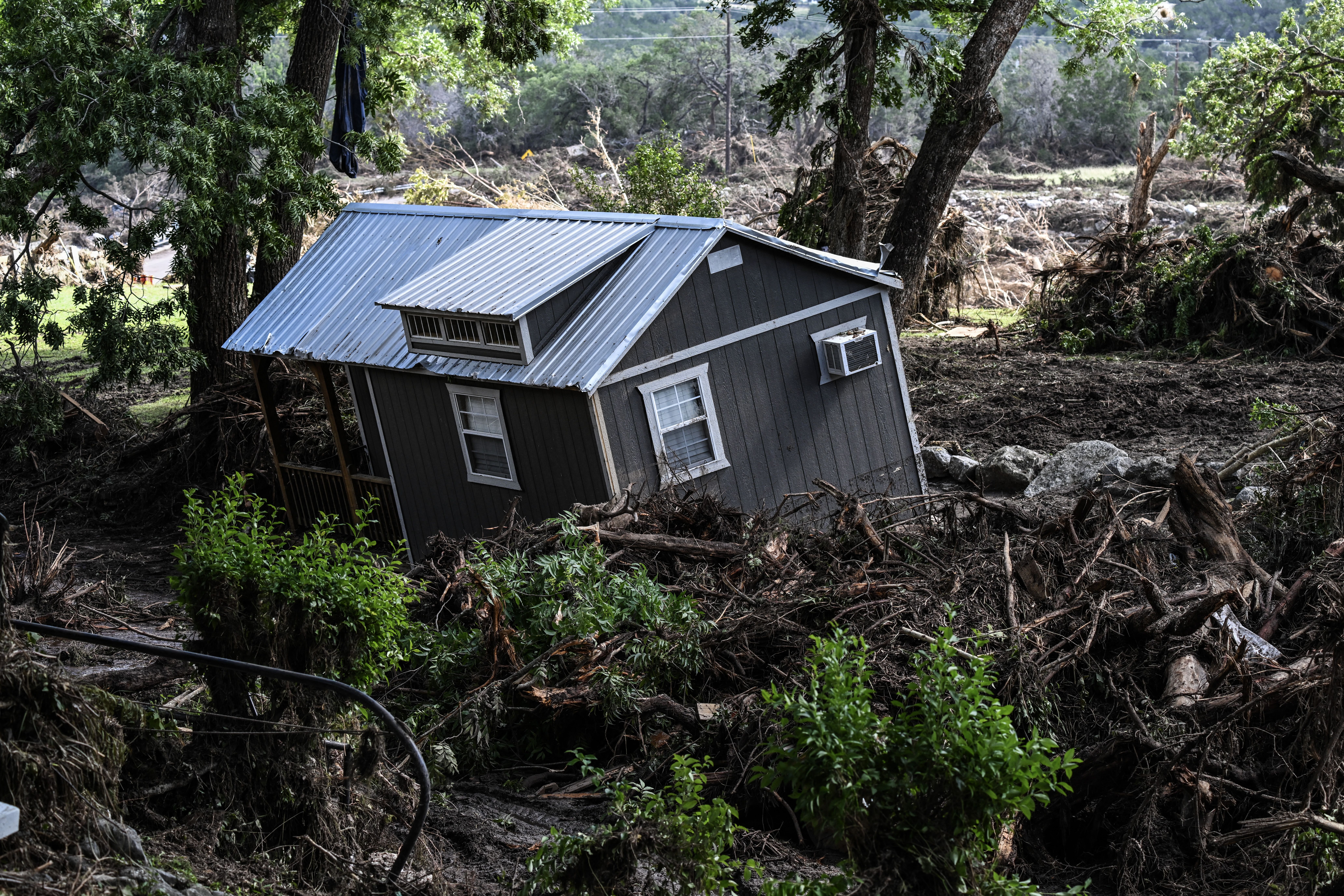 A damaged house is seen near the Guadalupe River in Hunt on July 8, 2025, following severe flash flooding over the July Fourth holiday. The small home is tilted into the ground and surrounded by fallen trees and debris.