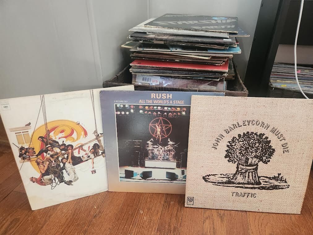 Three vinyl record covers on wooden floor: album with people on a swing, Rush's "All The World's A Stage" live stage photo, and Traffic's "John Barleycorn Must Die" with wheat bundle art.