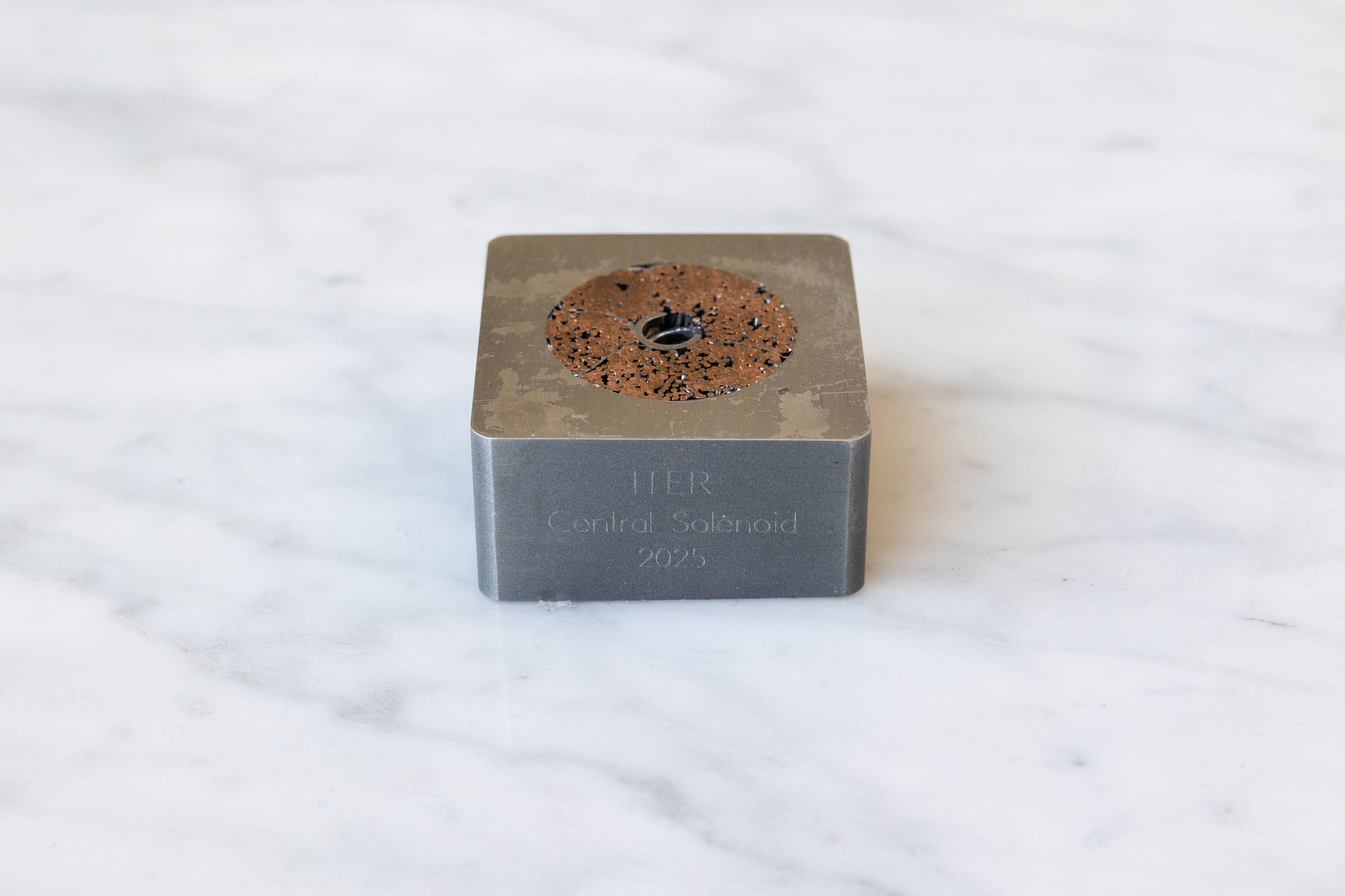 Grey square metal block on a white marble surface with a rusty copper ring embedded in the top, a central hole, and scattered dark specks; a faint inscription on the side reads Central Solenoid 2025.