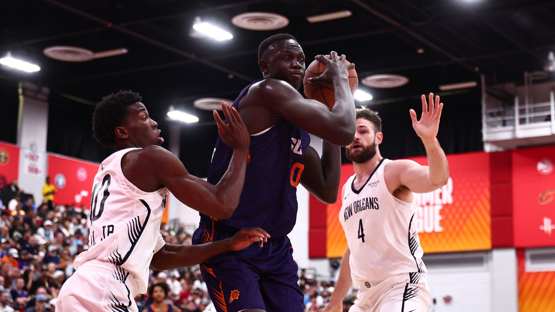 A basketball player in a purple jersey with orange and write writing on the front holds the ball after pulling down a rebound, while two players in white uniforms with black lettering defend him; fans are seated in bleachers in the background.