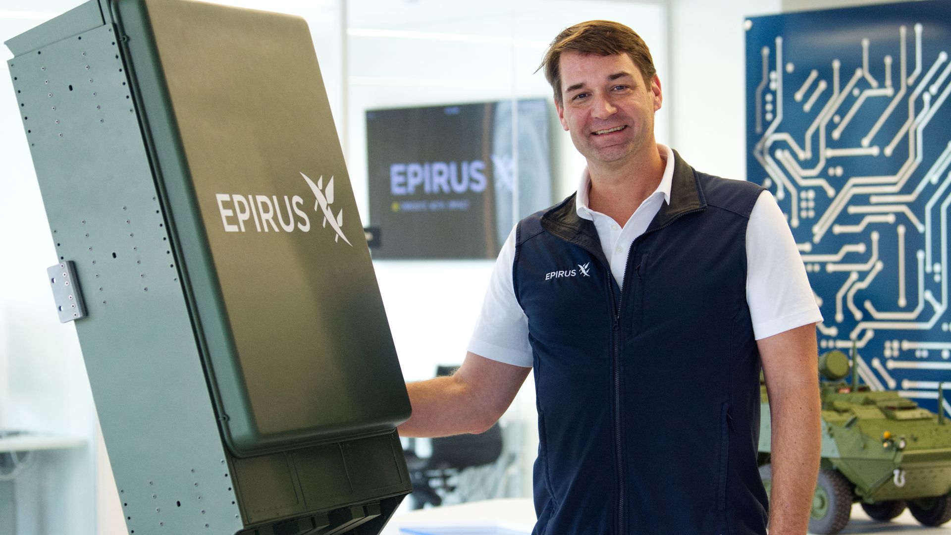 A man in a white shirt and blue vest stands next to a large directed-energy weapon model dubbed Leonidas. The background is white.