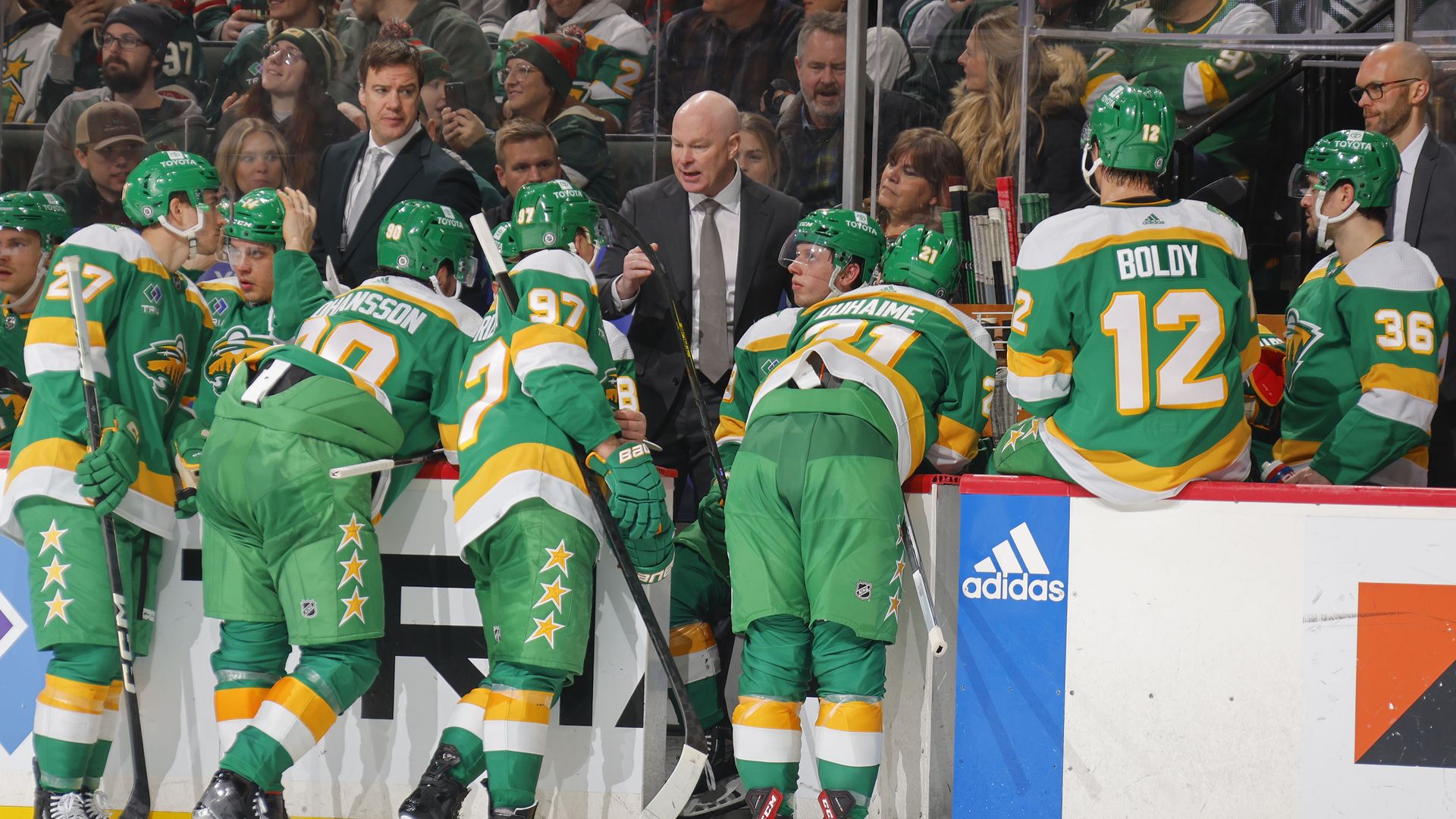 A group of hockey players dressed in bright green and yellow uniforms with large white numbers huddle near the white boards of a hockey rink next to a coach wearing a dark suit, who's pointing and giving instructions.