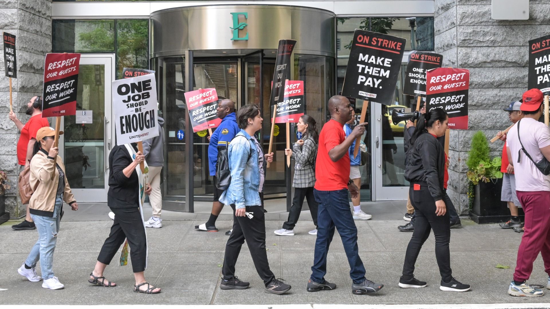 Group of diverse protesters marching outside a stone building, carrying red and black signs that read "ON STRIKE MAKE THEM PAY" and "RESPECT OUR GUESTS RESPECT OUR WORK"; some wear red shirts.