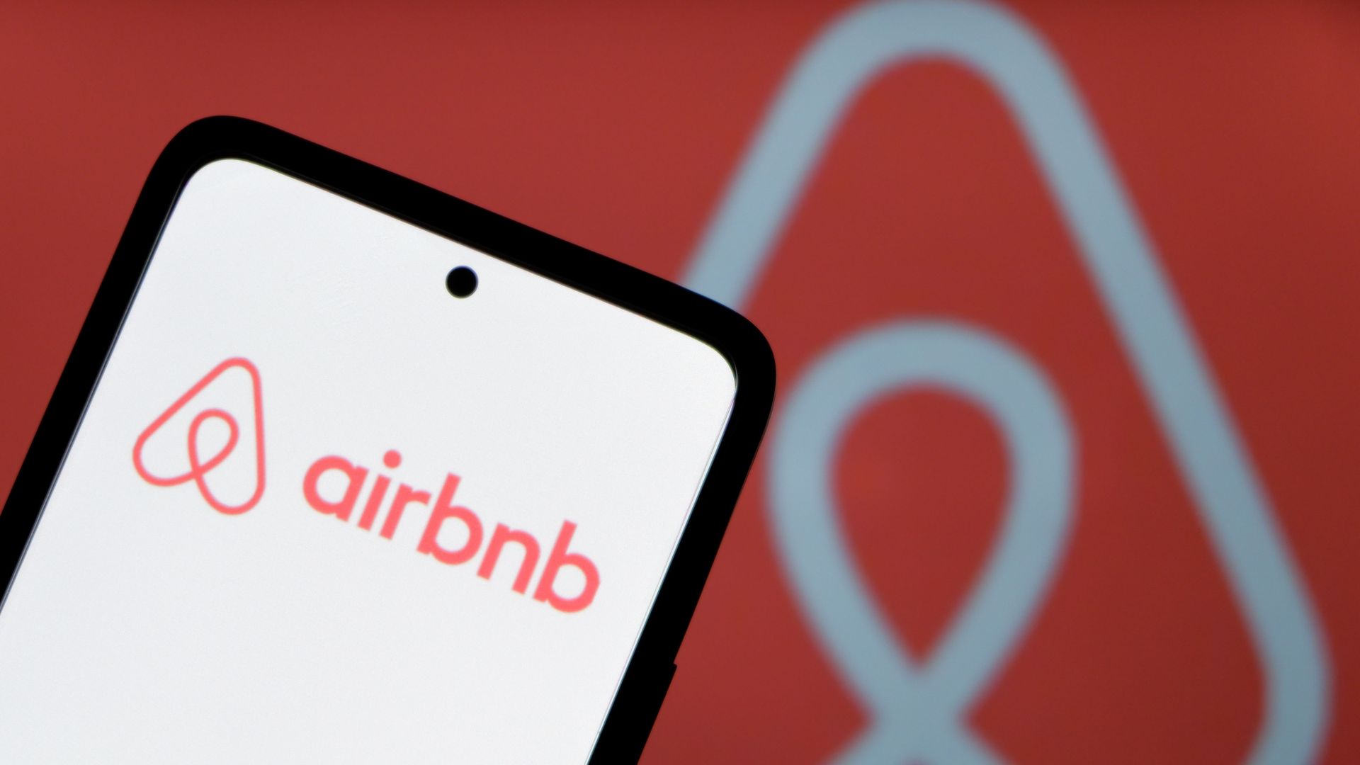 Close-up of a smartphone screen showing the word "airbnb" in pink/red on white, with a black edge; a large white Airbnb symbol appears on a red background in the backdrop.