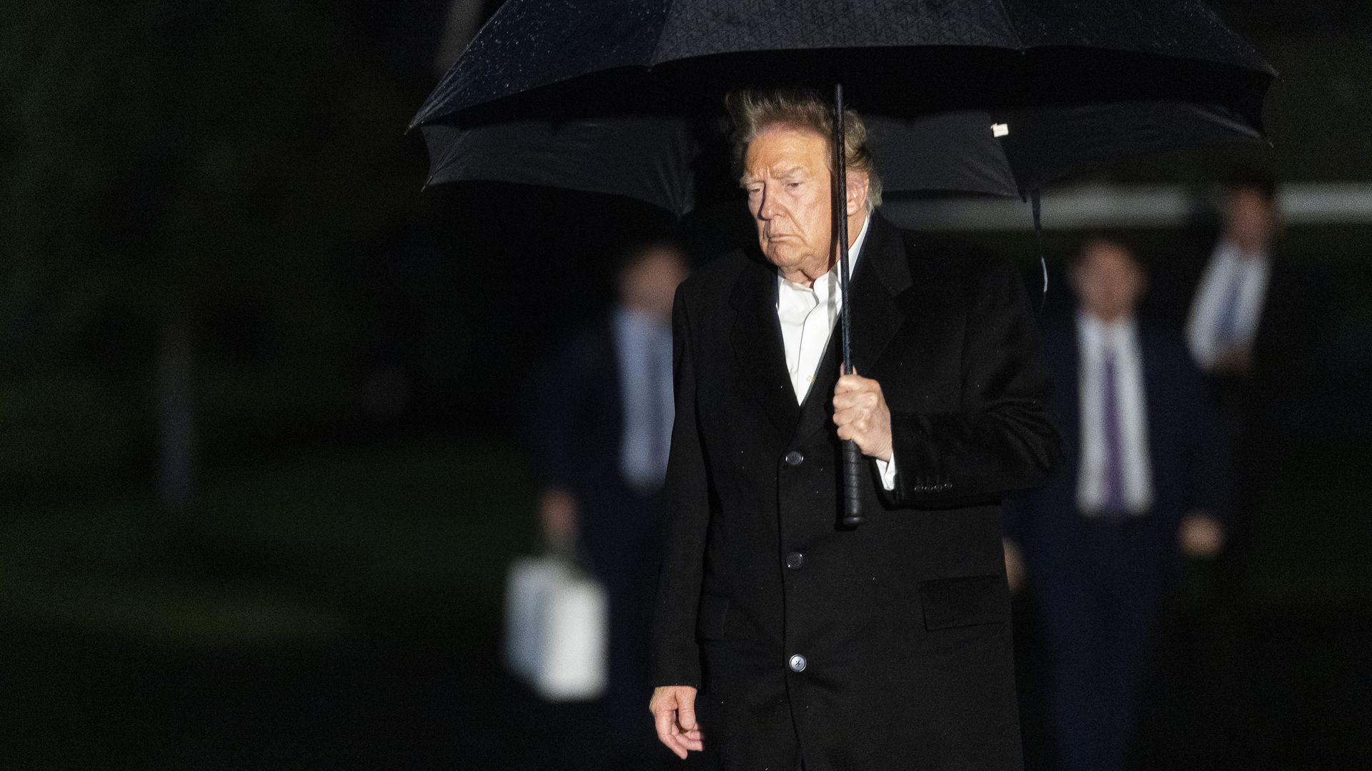 An older man, President Trump, in a black overcoat and white shirt stands in the rain under a large black umbrella, looking down with a serious expression, surrounded by dark, blurred figures.