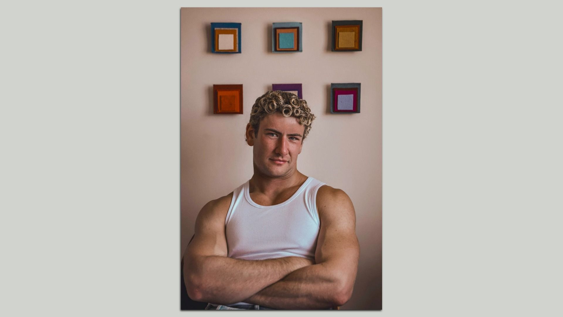 Muscular man with curly blond hair wearing a white tank top, sitting with arms crossed in front of a beige wall with colorful square artwork arranged in two rows.