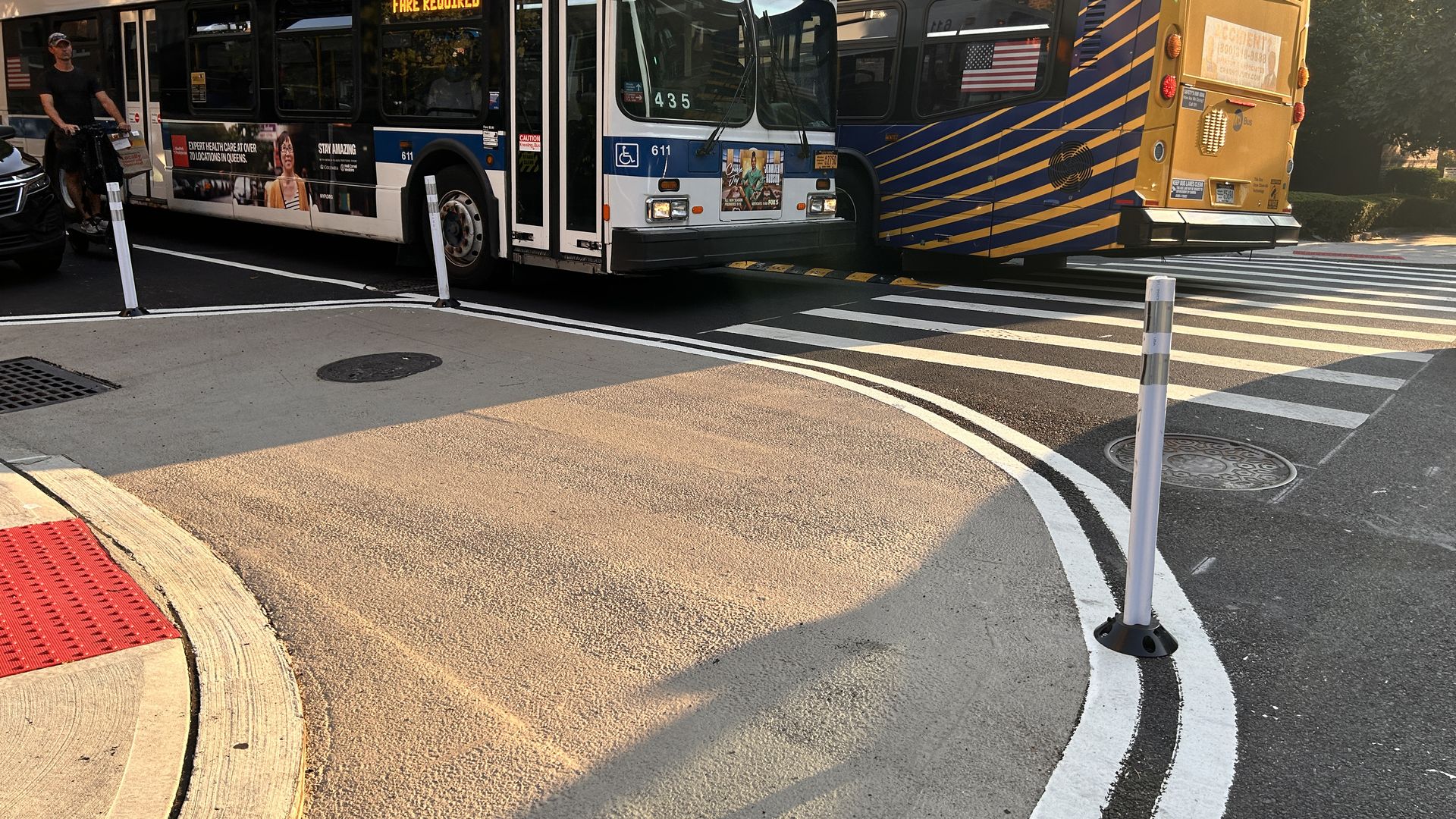 Two city buses: a white bus with blue trim and the "FARE REQUIRED" sign, and a blue-and-yellow striped bus, nose to tail at a crosswalk on a sunny urban street; a man stands by the white bus.