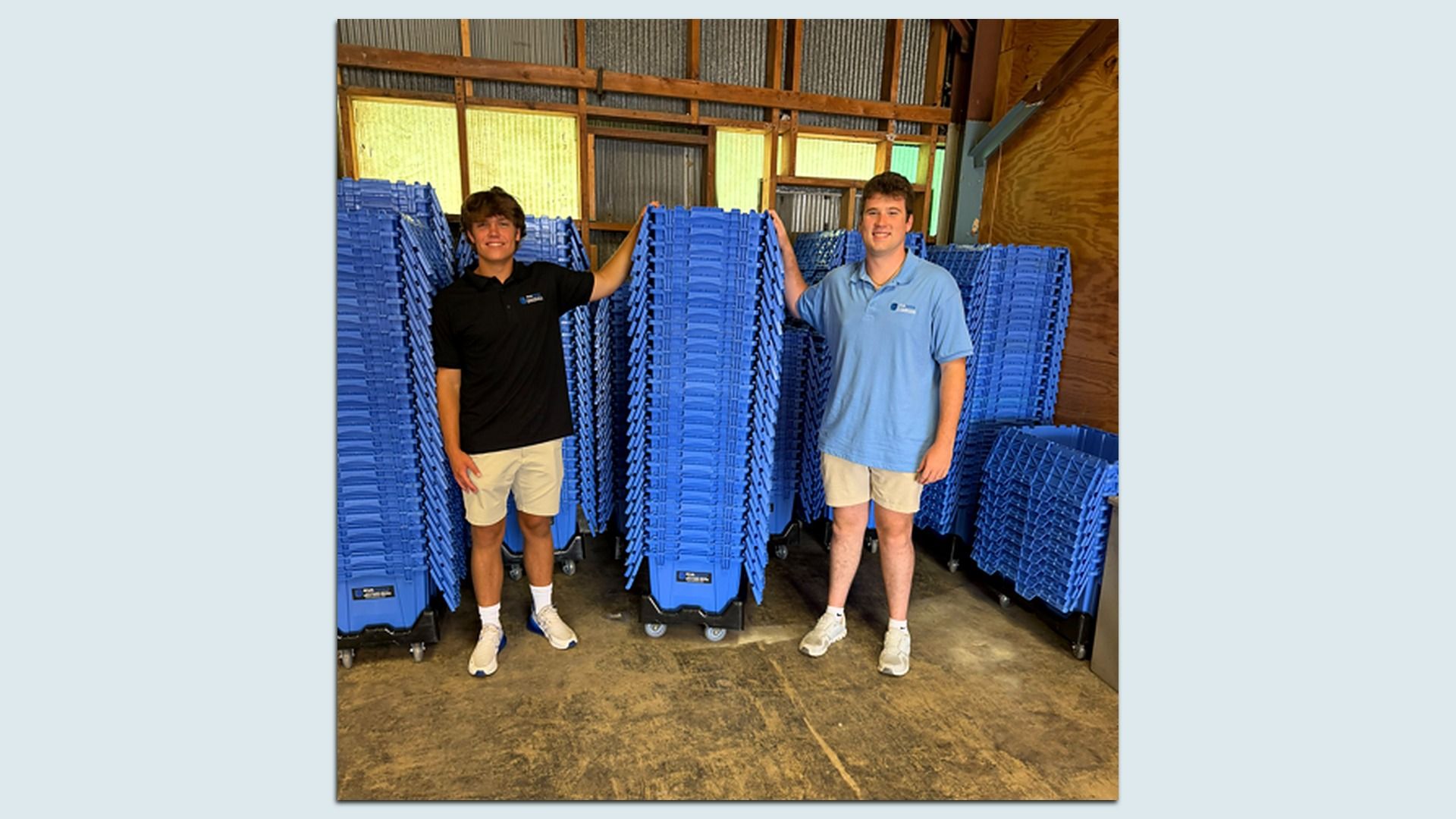 Two young men in polo shirts stand in a warehouse, smiling, leaning on tall stacks of blue plastic rolling crates, with wooden walls and metal siding in the background.