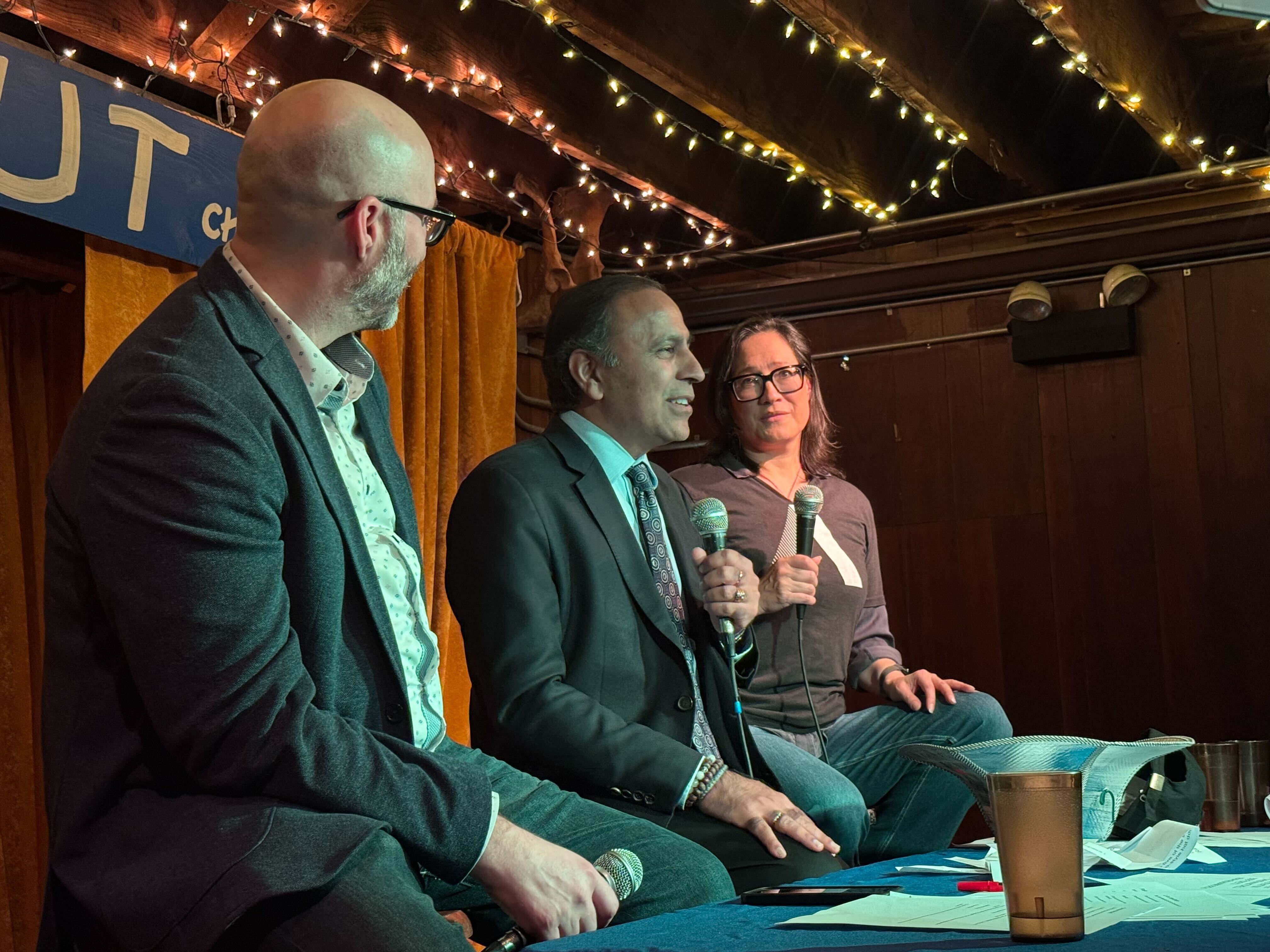 Three panelists sit on stage with microphones: a bald man in a blazer left, a suited man speaking middle, and a woman with glasses right. String lights and orange curtains behind.