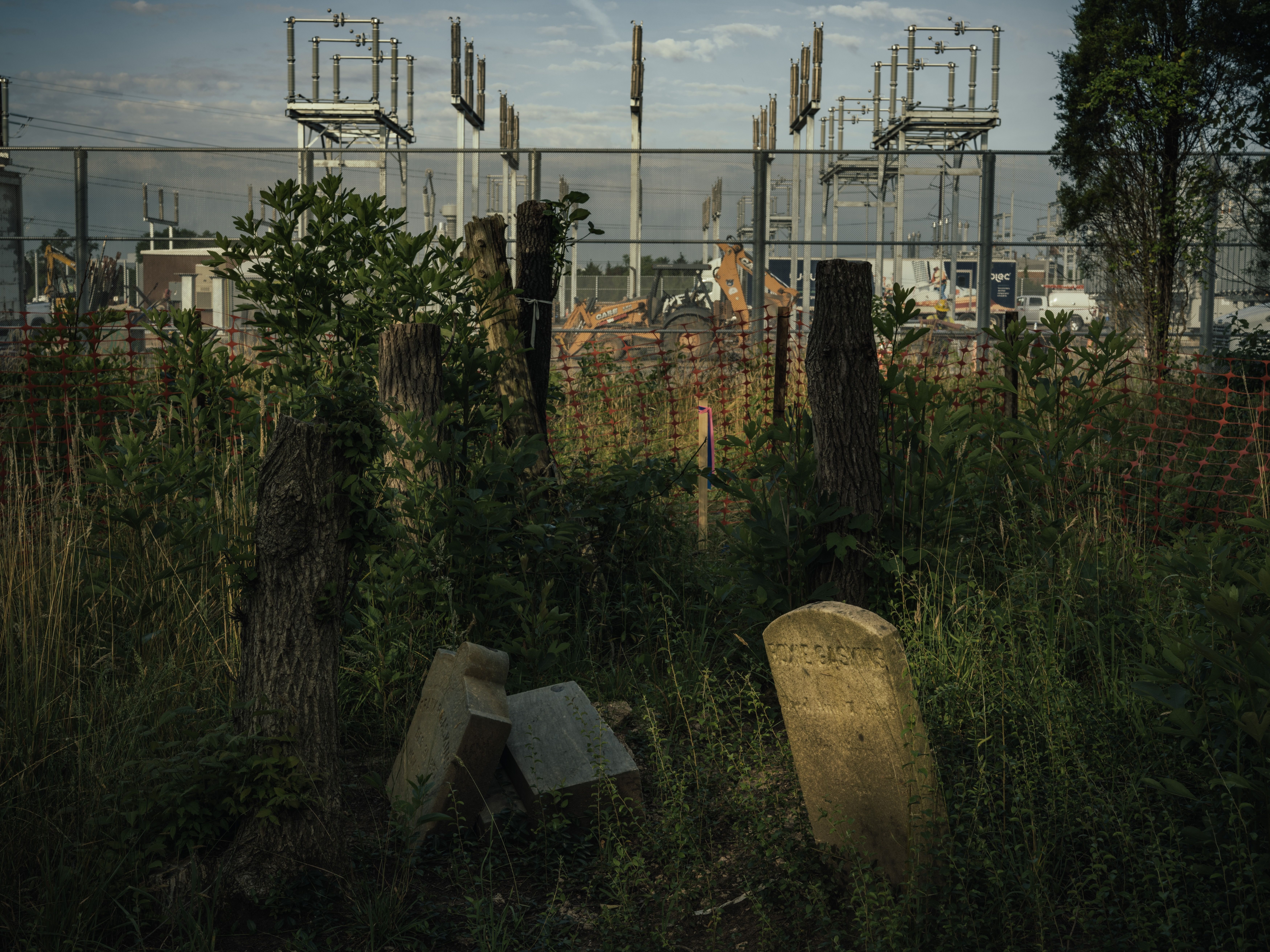 Overgrown foreground with shrubs and gravestones by a chain-link fence, overlooking a gray electrical substation with tall metal structures and construction machinery.
