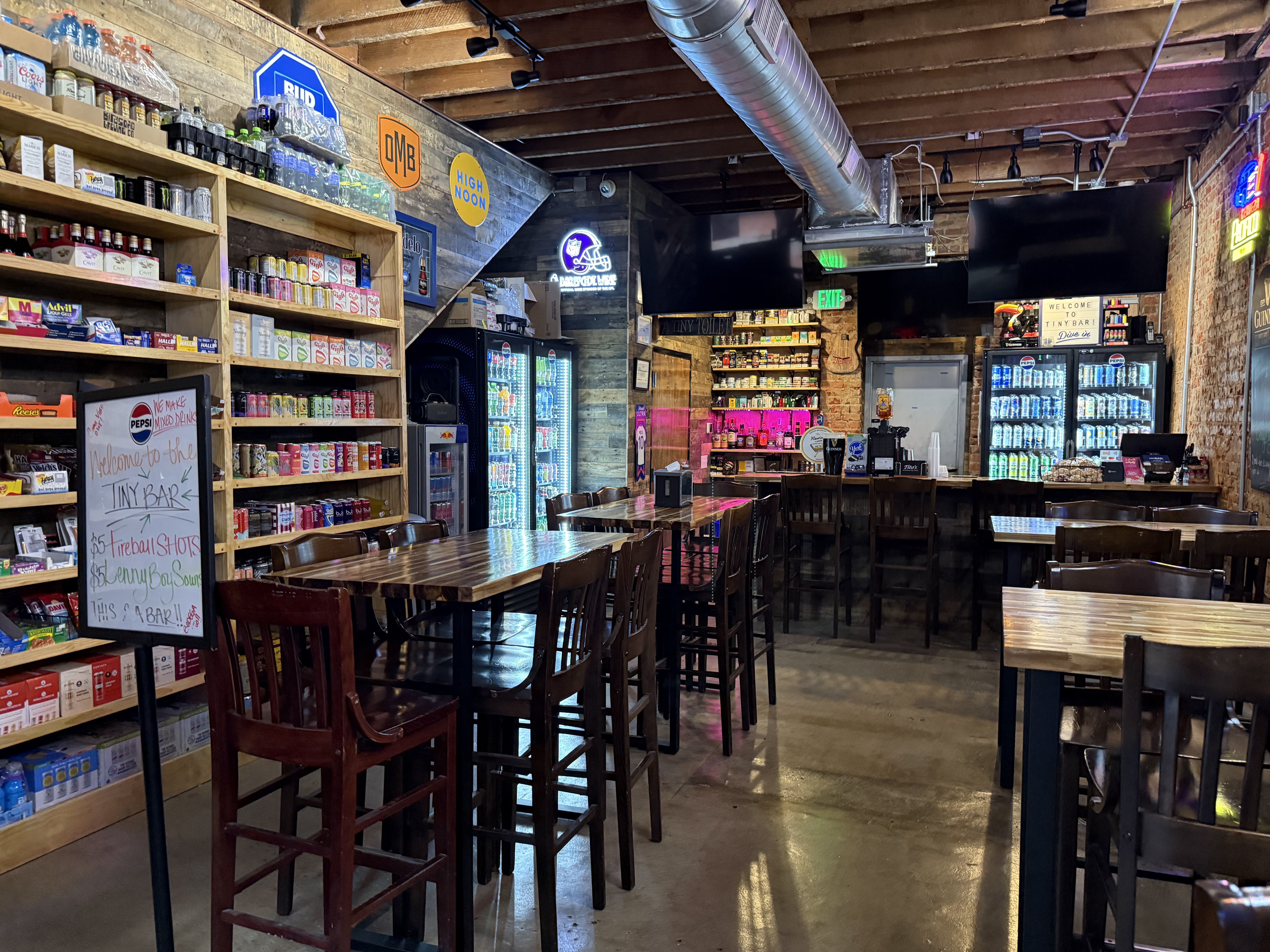 Interior of a rustic bar with wooden tables and chairs, shelves stocked with drinks and snacks, neon signs, and refrigerators filled with beverages under exposed wooden beams.
