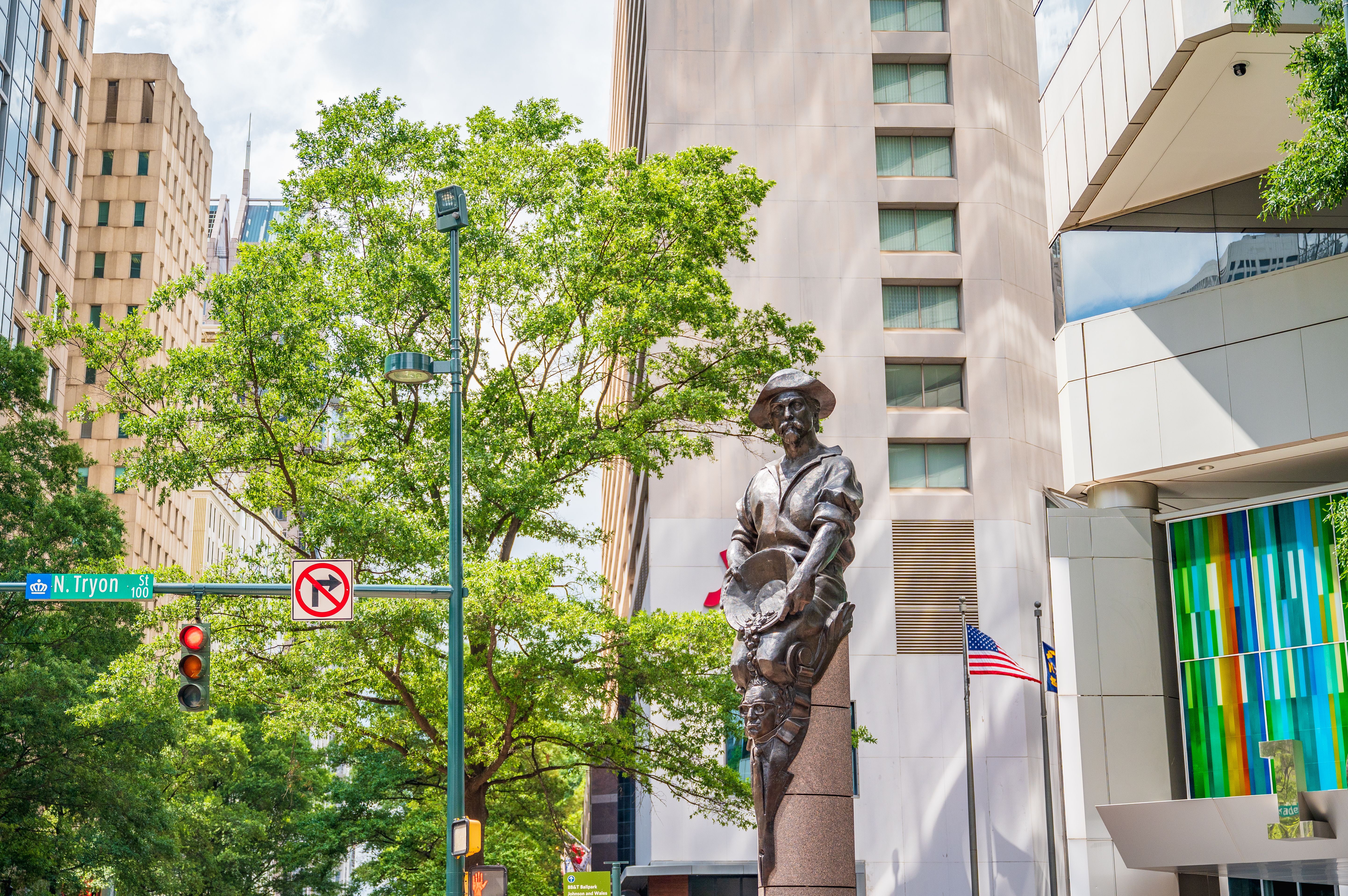 Sunny city street with green trees and modern beige and glass buildings, a bronze statue of a hat-wearing man, street signs reading N. Tryon St, traffic lights, and an American flag.