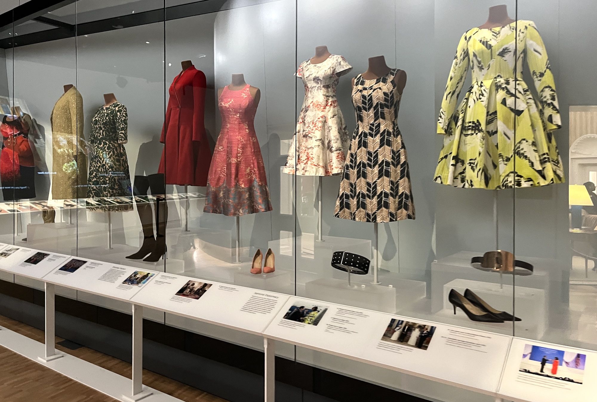 Window display with mannequins in colorful dresses: gold sequined coat, green floral, red, pink floral, white floral, beige-black geometric, and a yellow patterned cape behind glass.