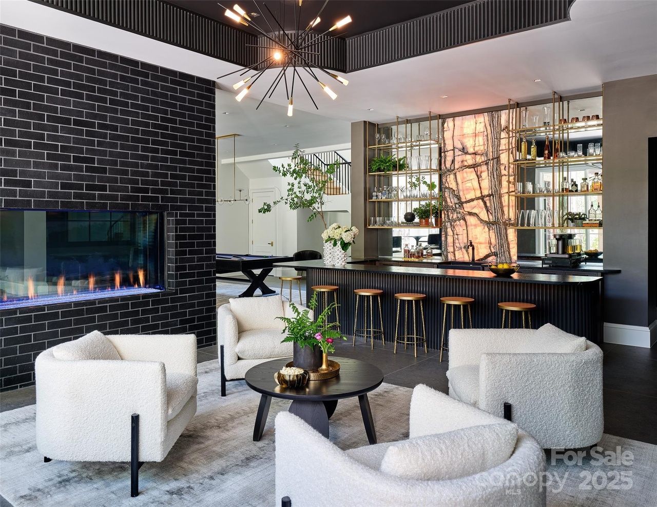 Modern living room with four white armchairs around a round black coffee table, black brick fireplace, and a stylish bar with gold stools and illuminated marble backdrop.